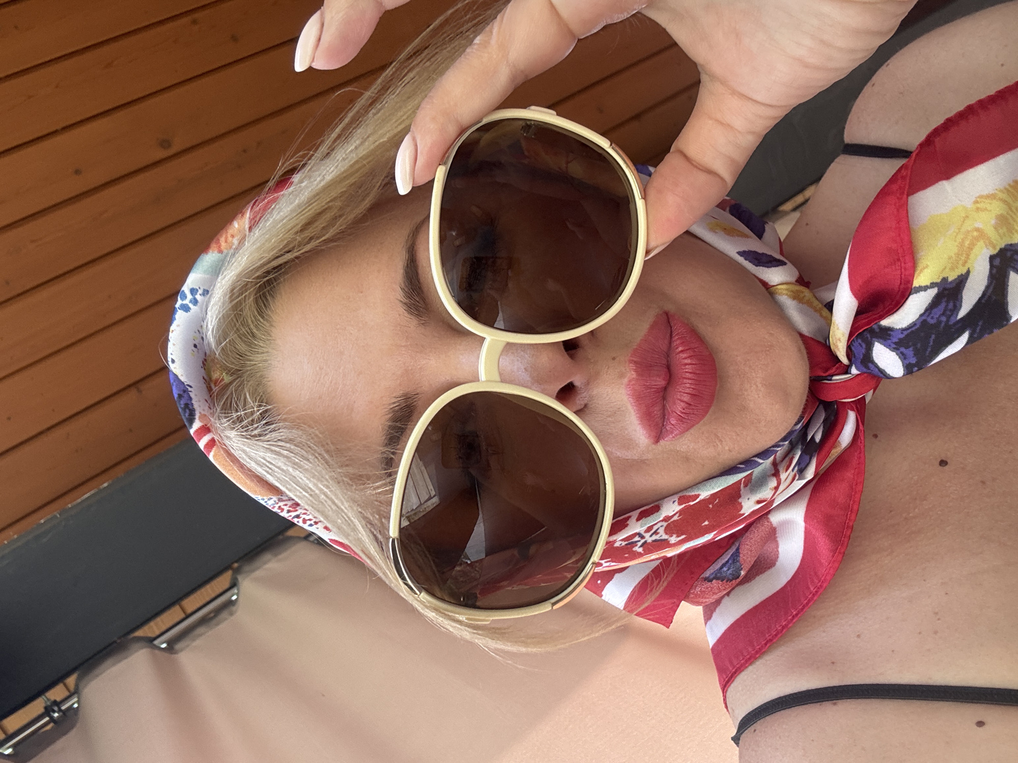 A medium shot captures a woman's face as she holds a pair of oversized, light-colored framed sunglasses to her face. She is wearing a colorful headscarf with red, white, yellow, and blue patterns. Her lips are painted a bright pink, and she has blonde hair. The background consists of wooden slats, suggesting an outdoor setting with a wooden ceiling or wall. The overall impression is that of a candid portrait taken in a warm, sunny environment, possibly outdoors in Sochi, Russia.