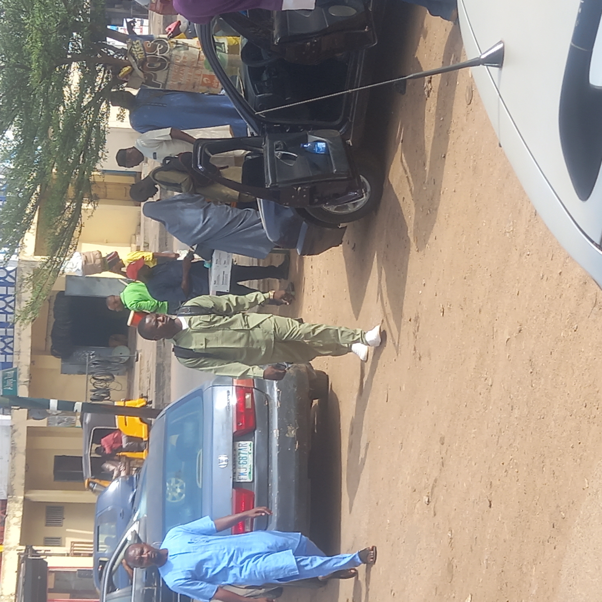 The image depicts a street scene in Dadi Riba, Nigeria. Several people are present, including a man in a light green uniform walking along the side of a parked grey Honda car. A man in a blue outfit stands near another grey car. Other people are visible in the background near a tricycle taxi. A white car is on the far right. Buildings line the street. The ground is covered with dirt and sand.