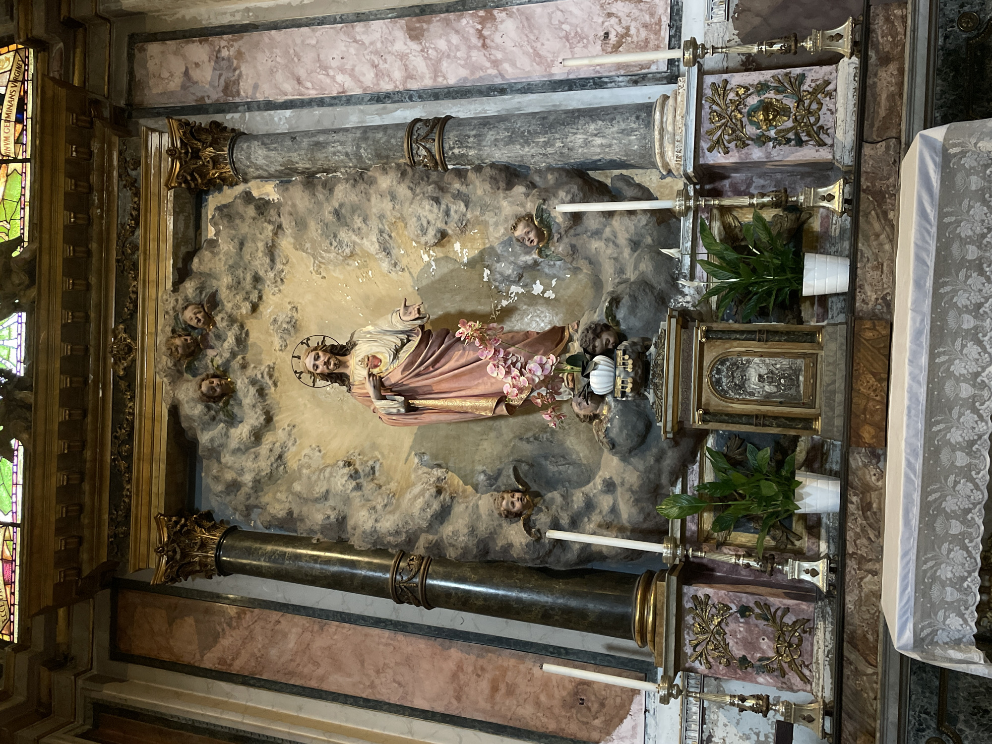 The image shows a decorated altar in what appears to be a Catholic church. The central feature is a three-dimensional relief depicting Jesus Christ lying down amidst clouds. Several cherubic faces are sculpted around him. Two dark marble columns with ornate gilded bases and capitals flank the relief. There are two candle holders with candles placed on the left and right sides. Two potted plants stand on the right, placed on the two decorated surfaces. A white lace cloth drapes to the right of the structure. Stained glass is visible on the left side. The location is possibly in Milan, Italy, given the architectural style and religious iconography typical of Italian churches.