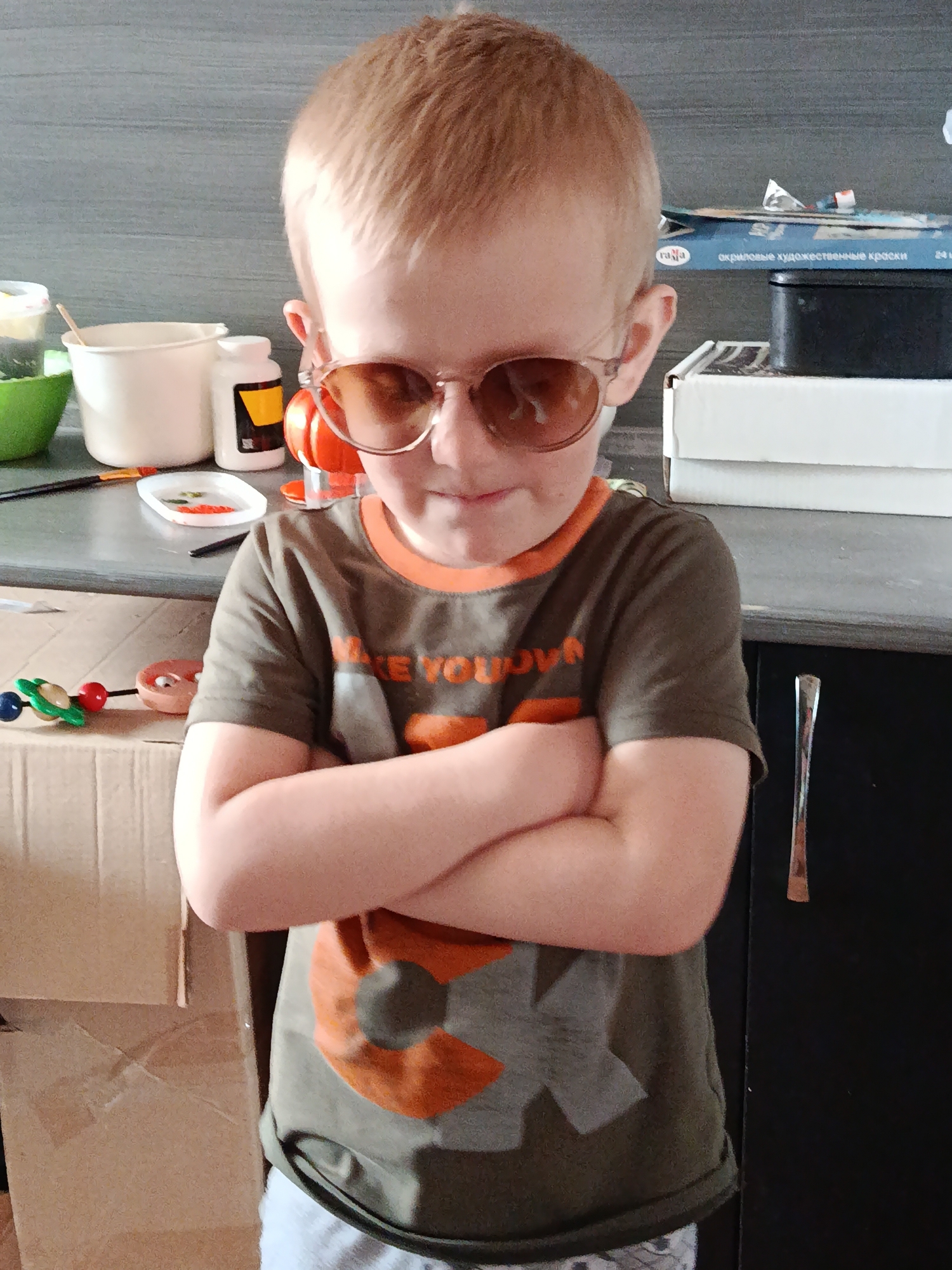 A young boy stands indoors with his arms crossed, wearing sunglasses and a t-shirt. Behind him, there's a table with craft supplies, a box, and a cabinet. The scene does not appear to be in Фоки, Russia; there's no evidence in the image to suggest that location.