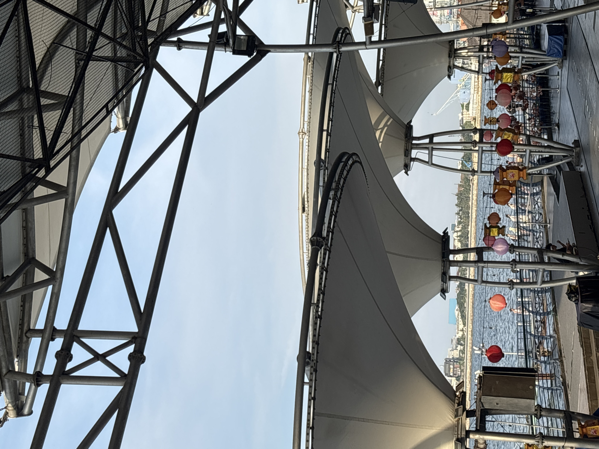 The image presents a view under a large, fabric-covered structure supported by a steel frame. The location is in North Jakarta, Indonesia, as evidenced by the cityscape and waterway visible through the structure. The framework is complex, with interconnected beams and supports. Strings of colorful lanterns and decorative objects hang from the structure. In the distance, there is a view of a city skyline along a waterway, with various buildings and structures visible. The sky is pale blue.