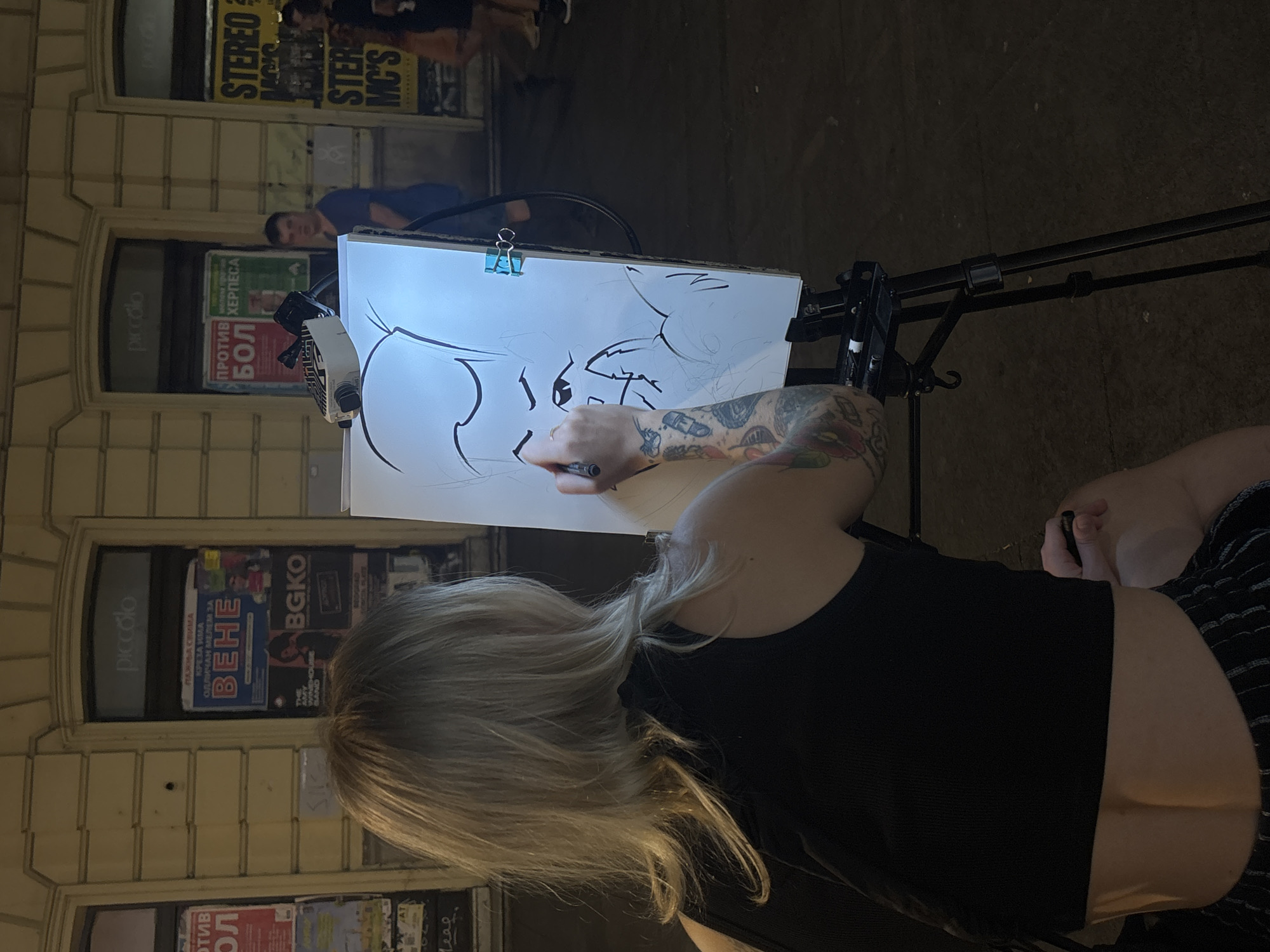 A woman with blonde hair and tattoos on her arms is drawing a caricature on a whiteboard. The whiteboard is on an easel. The woman is wearing a black tank top and striped shorts. She is holding a black marker in her right hand. In the background, there are buildings with signs. One sign says "piccolo". Another sign has Cyrillic text. There are people walking in the background. The scene appears to be on a street in Belgrade, Serbia.