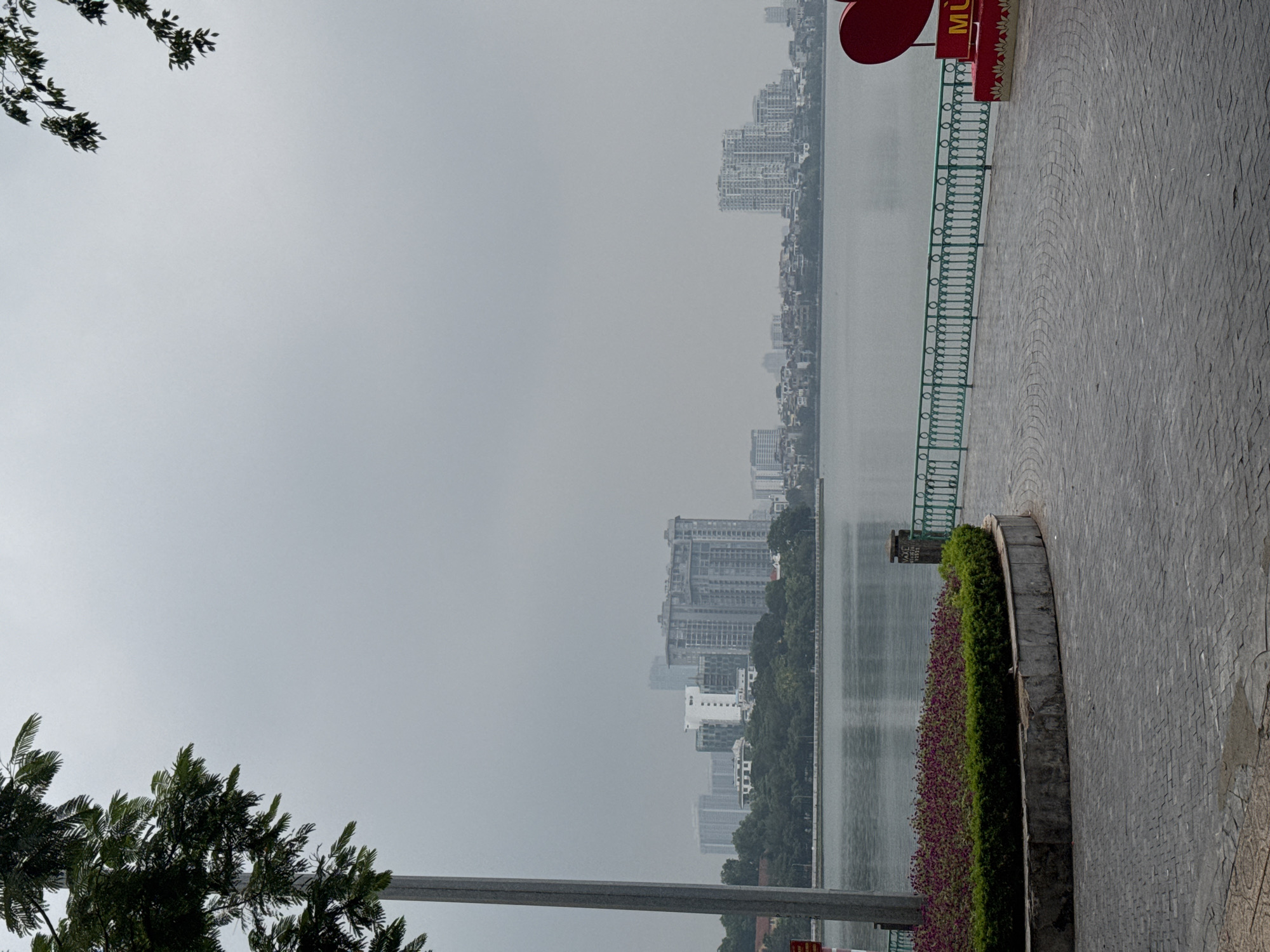 This is an outdoor, overcast vertical shot of a scenic city view in Hanoi, Vietnam. In the foreground, a paved walkway made of grey interlocking stones leads the eye toward a curved garden bed filled with small purple flowers and lush green bushes. A green metal railing borders the walkway along the water. To the right of the walkway, a large red heart sculpture with a red base and a yellow sign that reads "MU" is visible. In the midground, a wide body of water, likely a lake, reflects the hazy sky. Beyond the water, a dense cityscape of apartment buildings and other structures stretches across the horizon. Trees are scattered throughout the cityscape. A tall, grey lamppost stands on the left side of the frame, its top obscured by dark green foliage. The overall atmosphere is muted and tranquil, suggesting a peaceful, urban environment.