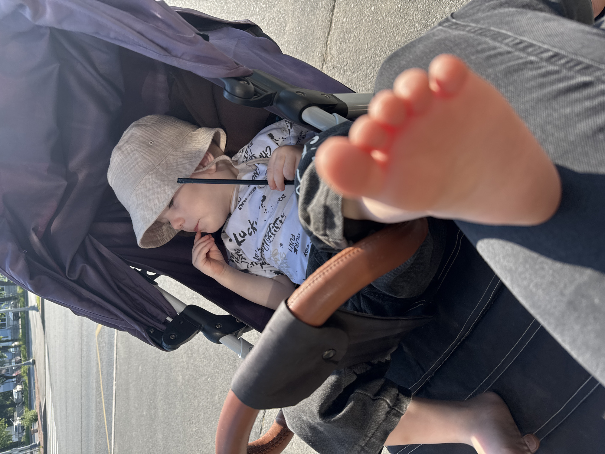 A young child wearing a bucket hat and a white t-shirt with black writing is sitting in a stroller. The child is holding a thin black object. The stroller has a brown leather-wrapped handle and a dark purple canopy. In the foreground, a bare foot is visible, with the toes extended. The child is wearing dark jeans. The background shows a street with a crosswalk, lampposts, and trees, suggesting an outdoor urban environment. The lighting indicates it is daytime, possibly late afternoon due to the angle of the shadows. The scene appears to be in a city, with evidence of infrastructure like roads and sidewalks.