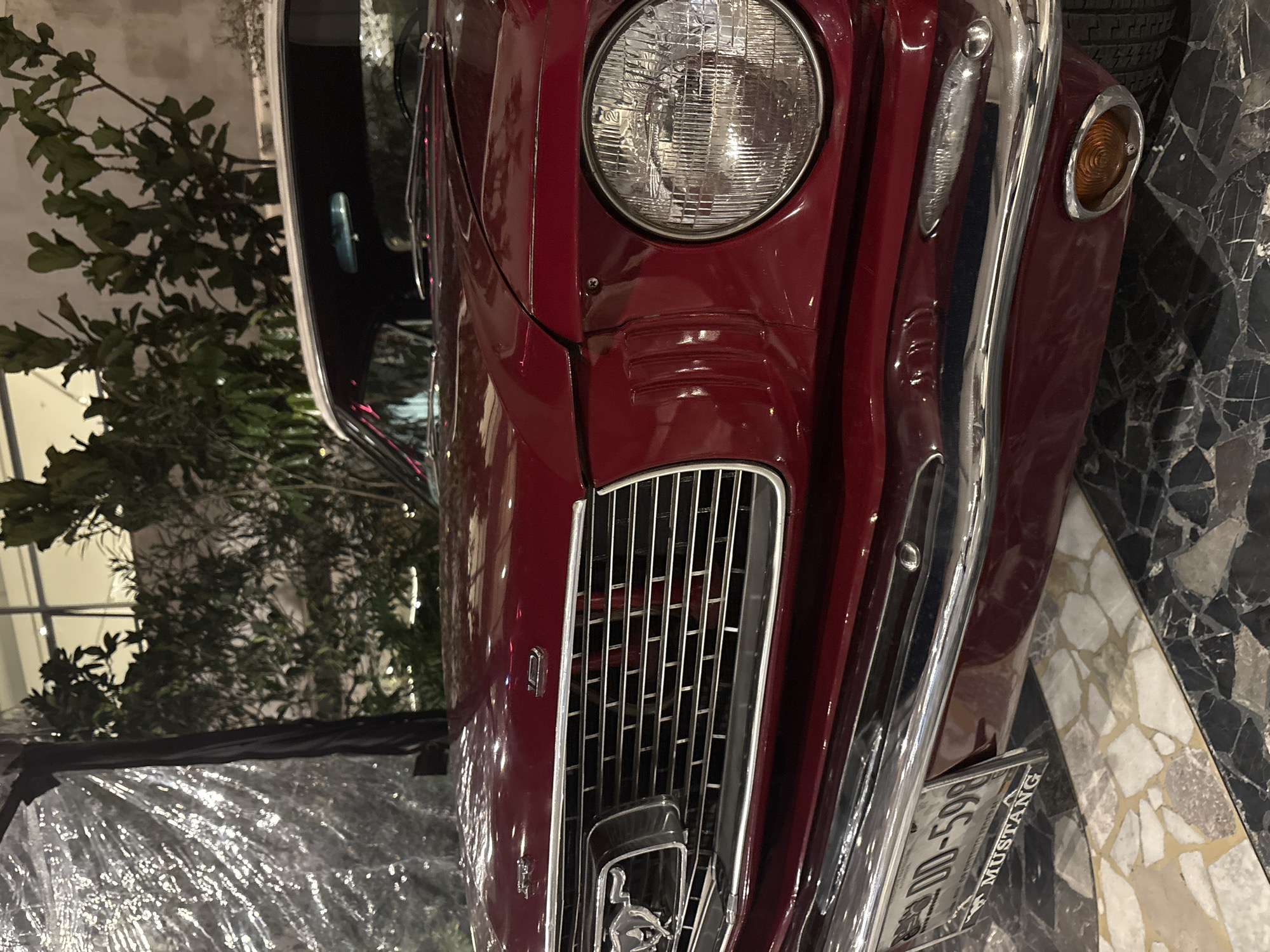 The image features a close-up view of the front of a red classic Mustang. The car’s grill, headlight, and chrome details are clearly visible. The car is parked in what appears to be an indoor location, with a marble-like tile floor. A plant can be seen in the background on the left. The license plate on the vehicle reads "DD-598" and includes the word "MUSTANG". The image was taken in Mexico City, Mexico.