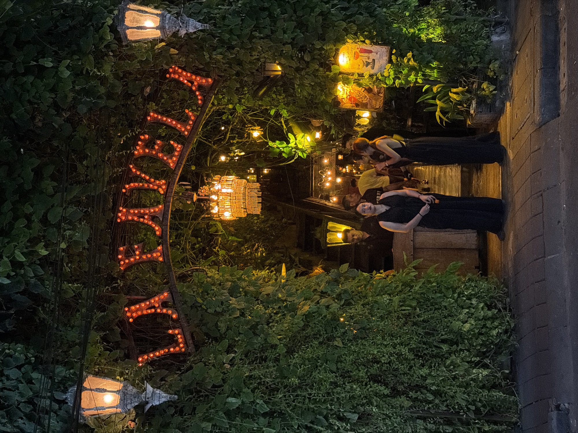 A nighttime photograph of the front of a restaurant called "La Favela" in Kuta, Indonesia. The restaurant’s entrance is framed by dense, green foliage, with two lanterns hanging on either side of an arched sign that spells out the restaurant's name in red lights. A crystal chandelier hangs further inside the entrance.
Three people are in the foreground. One person, dressed in a black dress, stands and faces the camera. Two other people stand behind her, partially obscured by greenery and lighting. They appear to be in conversation.
The entrance to the restaurant is to the right of the group, framed by greenery and leading towards interior seating and decor. The foreground is a sidewalk adjacent to the building, with a dark curb visible on the right side of the frame.