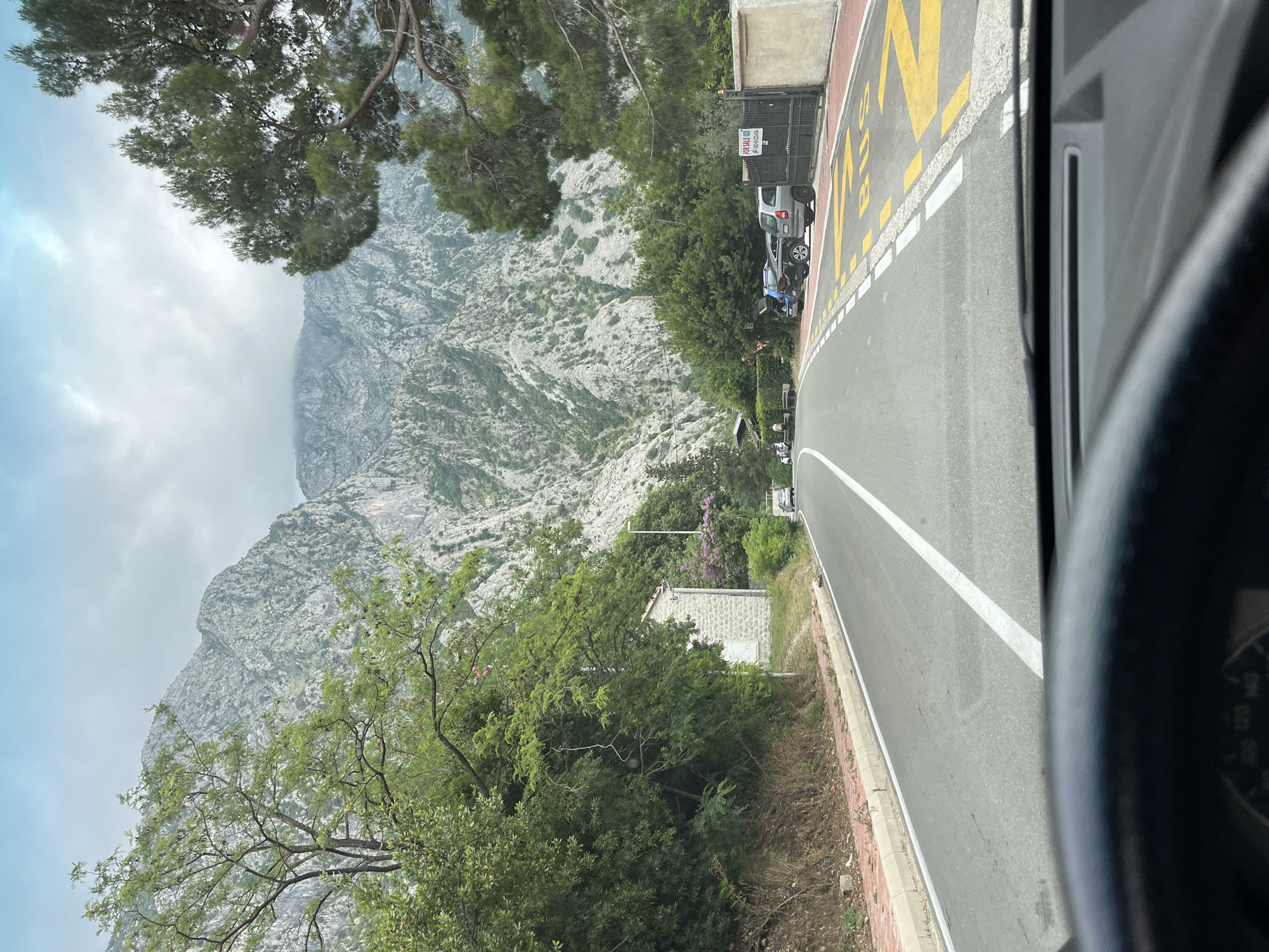 The image displays a scenic mountainous landscape, likely captured from inside a vehicle, with a distinct lean suggesting it was originally a portrait-oriented photograph presented here rotated ninety degrees clockwise.
In the foreground, the dark interior of a car, including part of the dashboard and a pillar, is visible on the far right edge. To the left of this, an asphalt road curves gently into the distance. It features a continuous white line along its inner edge and a dashed white line marking a lane or shoulder. Adjacent to the road, on the right, is a red-paved area with large yellow letters clearly spelling "BUS."
On this red shoulder, several vehicles are parked, including a silver SUV and a dark sedan. Next to the parked cars stands a black metal gate, bearing a white sign with prominent red text that reads "FOR SALE," followed by a partially visible phone number.
The mid-ground is filled with lush green trees and bushes, behind which a small, stone-built house with a white door is nestled. The background is dominated by a majestic, rugged mountain range composed of greyish-white rock, sparsely covered with green vegetation. The upper sections of the mountains are partially obscured by low, grey clouds or mist, indicating an overcast or partly cloudy day. The overall scene evokes a sense of traveling through a picturesque, natural environment in Donji Orahovac, Montenegro.