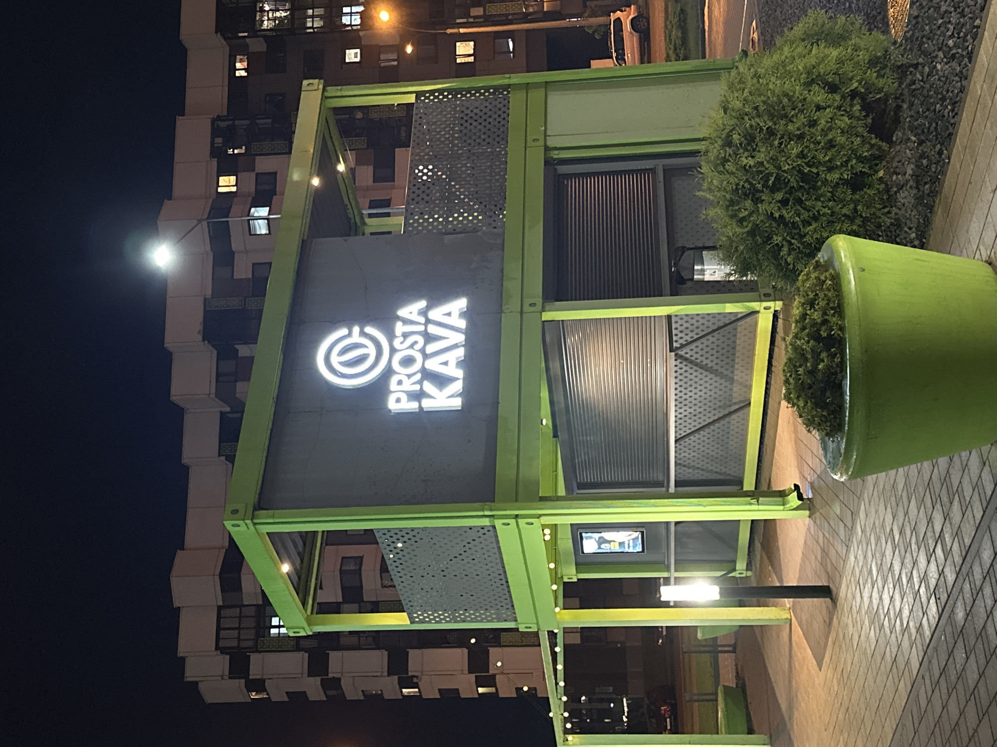 The image shows a Prosta Kava coffee shop in Бараўлянскі сельскі Савет, Belarus. The shop is located in a modern building with multiple floors and many windows. The shop itself is a green structure with the Prosta Kava logo prominently displayed. The entrance is visible with roller shutters. There is a large green planter with a bush next to the shop. The street is paved with stone tiles. It is nighttime, and the scene is lit by streetlights and the shop's lighting. There is a building visible in the background.
