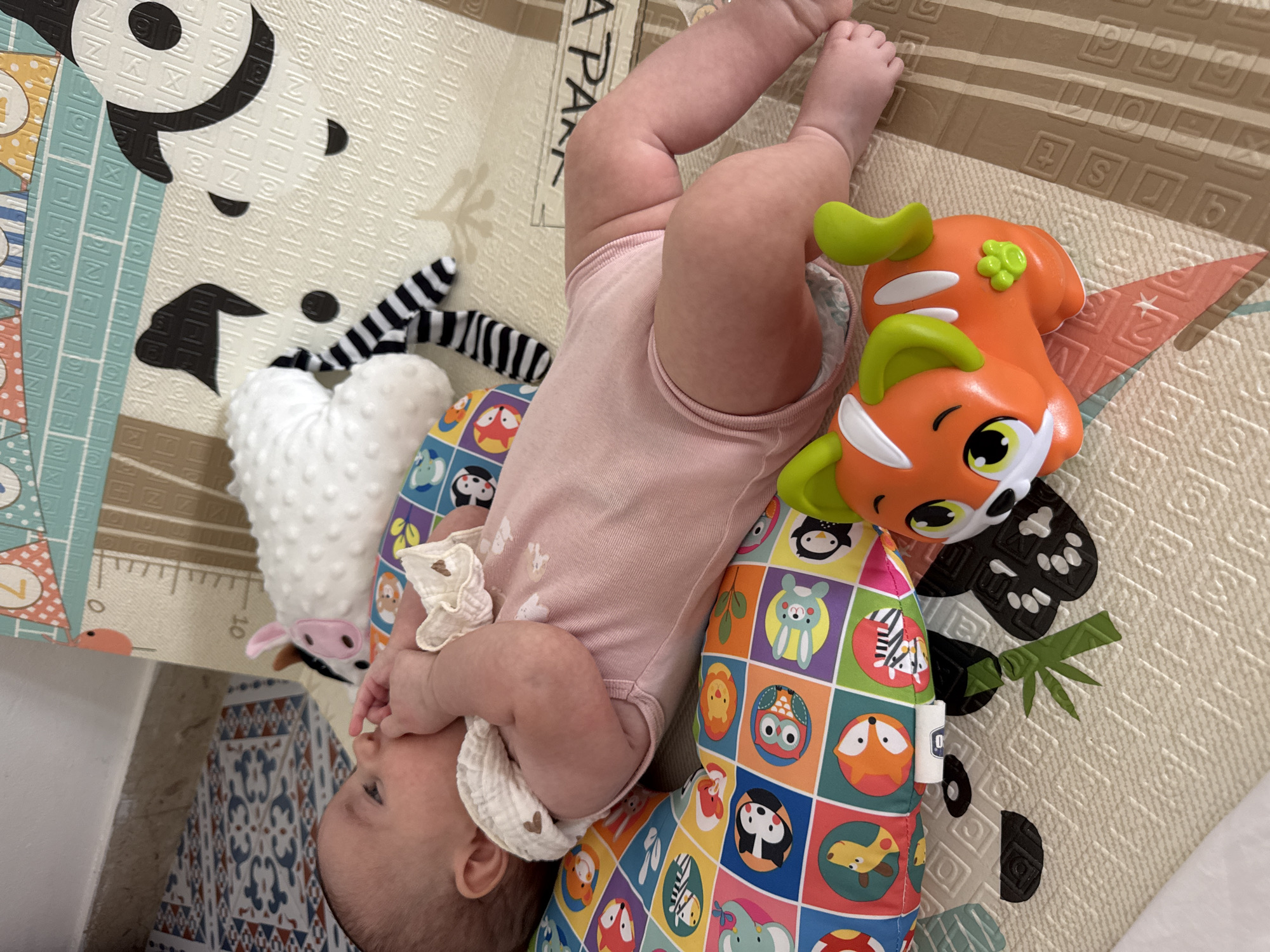 A baby is lying on its back on a colorful play mat. The baby is wearing a pink onesie and has its hands in its mouth. There are several toys on the play mat, including a stuffed animal that looks like a cow and an orange toy that resembles a dog. The play mat is positioned on a tiled floor.