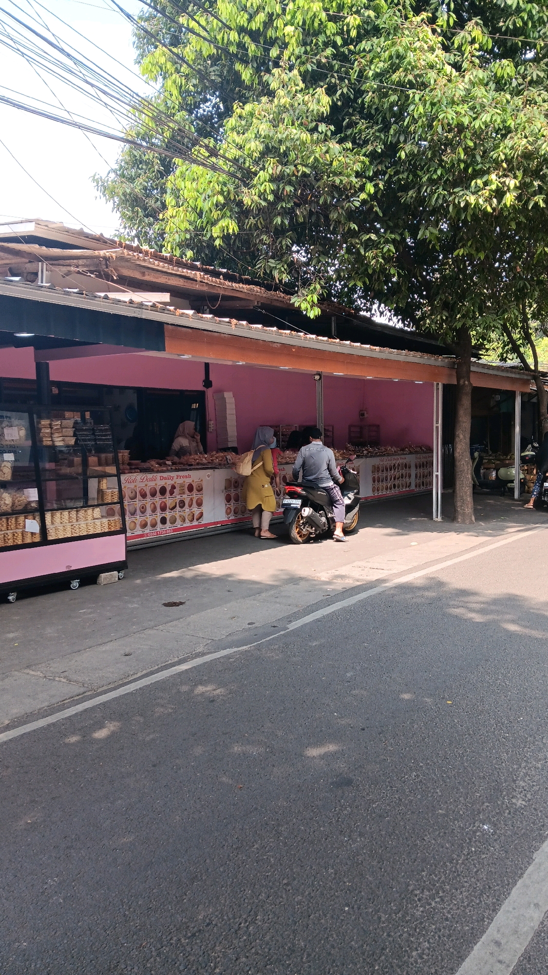 In South Jakarta, Indonesia, on a sunny day, a street-level view captures a bakery storefront with a bright pink facade and a brown, corrugated roof. To the left, a glass display case filled with various baked goods sits on wheels. In front of the counter, two individuals stand: one, a woman in a yellow dress and headscarf, faces the counter, while another, a man in a grey shirt and dark pants, sits astride a black motorcycle parked on the asphalt road. The motorcycle is positioned at an angle, facing away from the viewer. Behind them, a long counter displays more baked goods, and a sign reads "Roti Dalil Daily Fresh." Overhead, a dense canopy of green leaves from a large tree drapes across the scene, with a network of power lines crisscrossing the sky. The road in front of the bakery is asphalt, with a concrete sidewalk bordering it. Further down the street, other businesses and trees are visible.