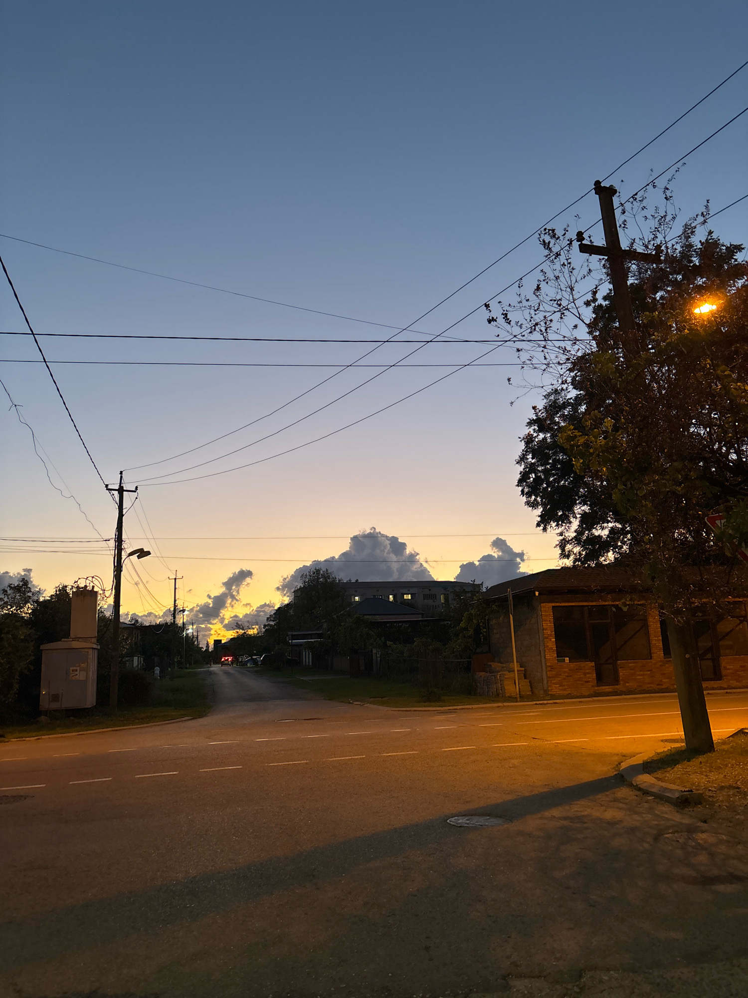 The image shows a street scene at dusk in an urban or suburban area. The sky is a gradient of blues and oranges, with fluffy white and grey clouds illuminated by the setting sun. The foreground is dominated by a paved road with white lane markings and a manhole cover. To the right, a tree casts a long shadow across the road, and a lamppost with an illuminated orange light is visible. A brick building with large window openings is also on the right side of the road. On the left, there is a utility box on a pole, and further down the road, power lines crisscross the sky. Trees and houses are visible on both sides of the road, and a few cars are in the distance. The overall impression is one of a quiet, end-of-day atmosphere.