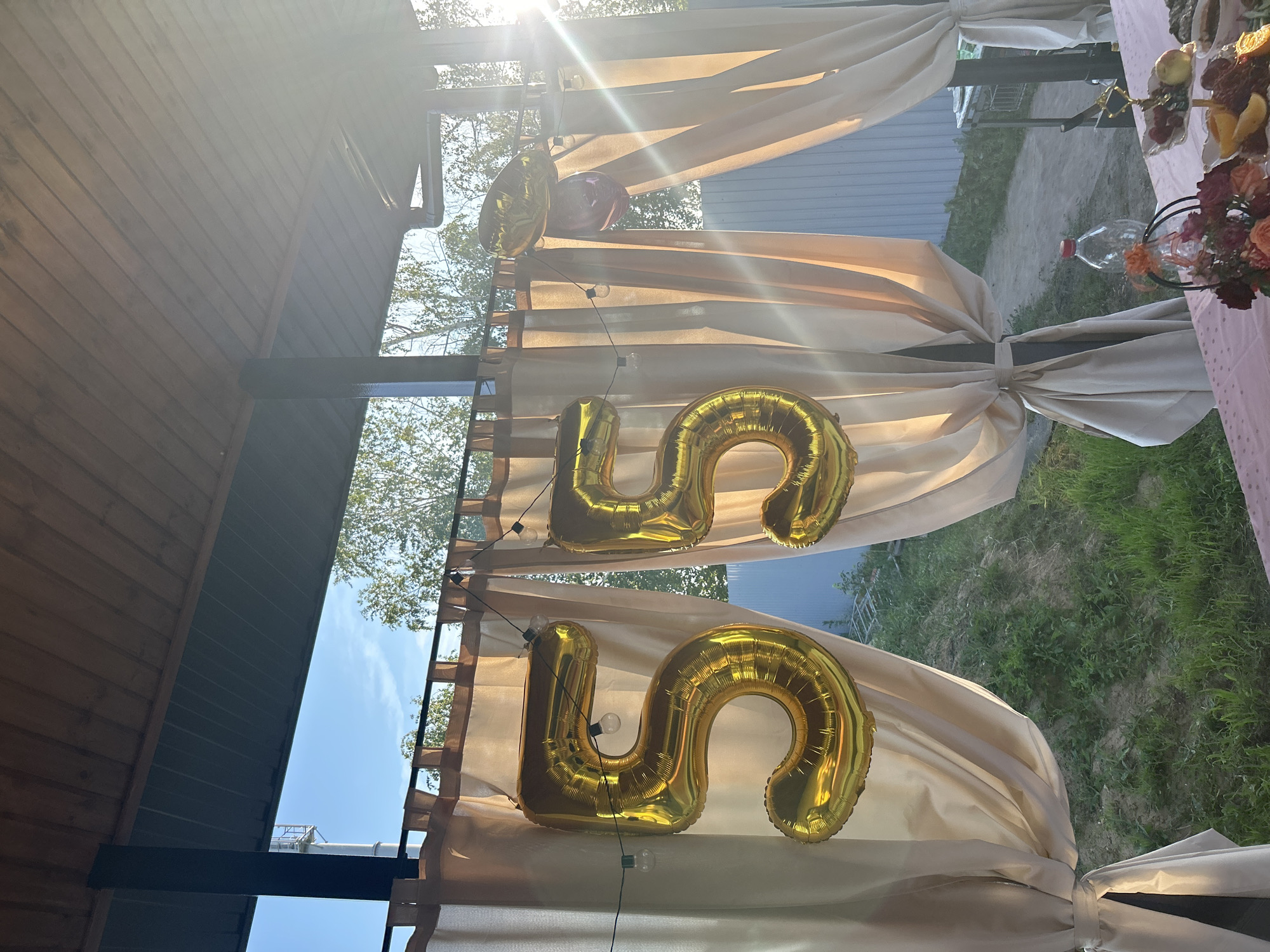 In Pribrezhnyi, Russia, a celebratory outdoor scene unfolds under a covered patio. Two large, golden balloons, shaped like the numbers "5" and "5," are prominently displayed, suspended by string lights. These balloons suggest a 55th birthday or anniversary celebration.
Behind the balloons, light beige curtains hang, framing the view of the outdoors. Through the gap in the curtains, a bright sunbeam cuts across the scene, creating lens flare and casting a warm glow. The background reveals lush green grass and trees, with a glimpse of a light blue sky. A light-colored metal fence is also visible in the distance.
The foreground features a pink tablecloth draped over a table, hinting at a prepared feast. A bouquet of flowers in shades of orange and deep red sits in a dark basket. Various dishes, including fruit and what appears to be charcuterie, are arranged on the table, ready for consumption. A clear plastic water bottle stands near the floral arrangement. The overall impression is one of an outdoor gathering, likely a birthday party, set in a suburban or dacha environment.