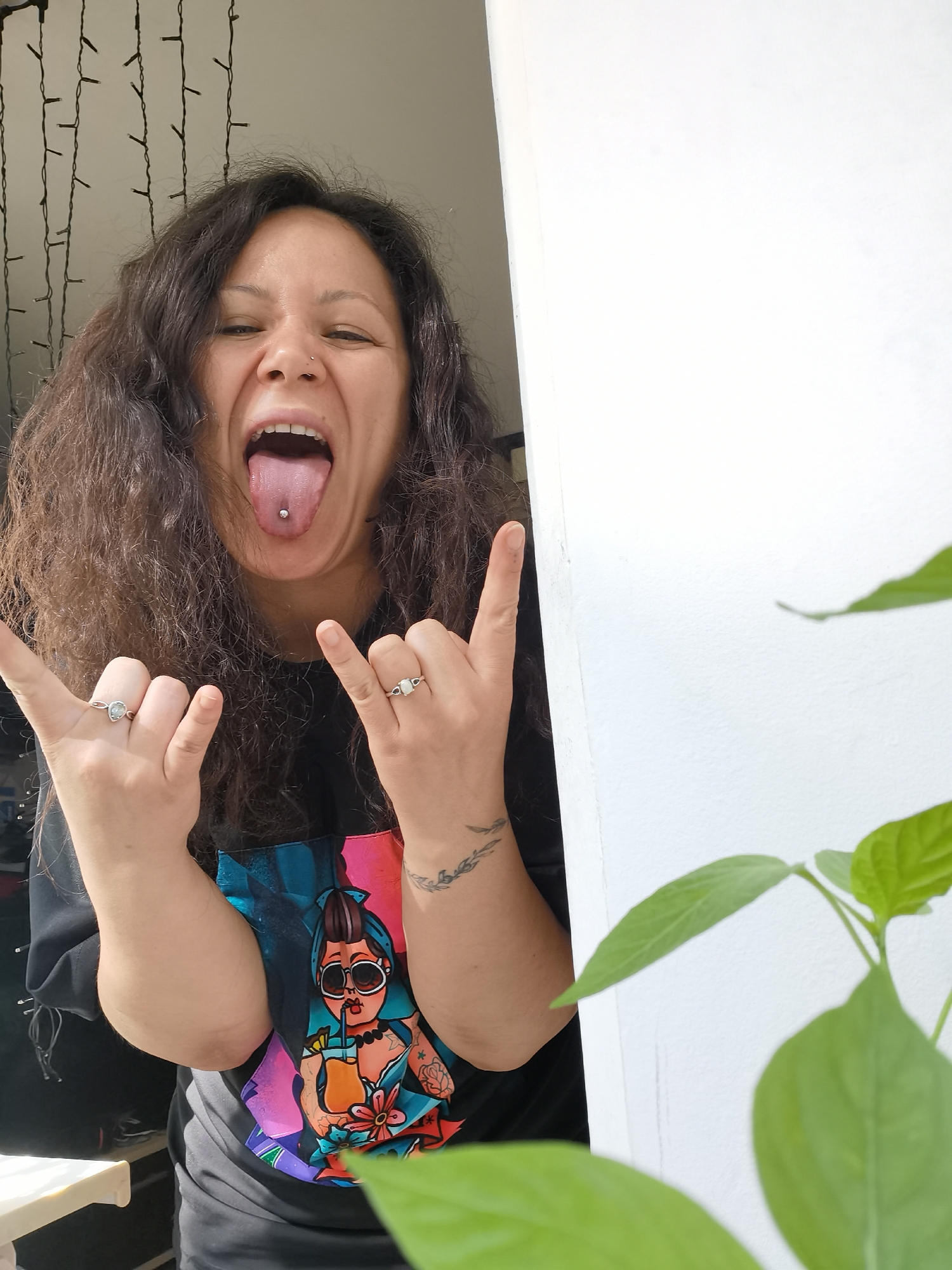 A medium shot of a person with long, dark, curly hair, piercing on their nose and tongue, and a tattoo on their left arm, striking a “rock on” hand gesture with both hands. They are wearing rings on both ring fingers and a black t-shirt with a colorful graphic of a woman with sunglasses and a drink. The person is standing in front of a white wall with green leaves of a plant visible on the right side of the frame. Behind the person, through a doorway, there are lights hanging in the background. The location is indicated as Chaikovsky, Russia, although this cannot be verified visually.
