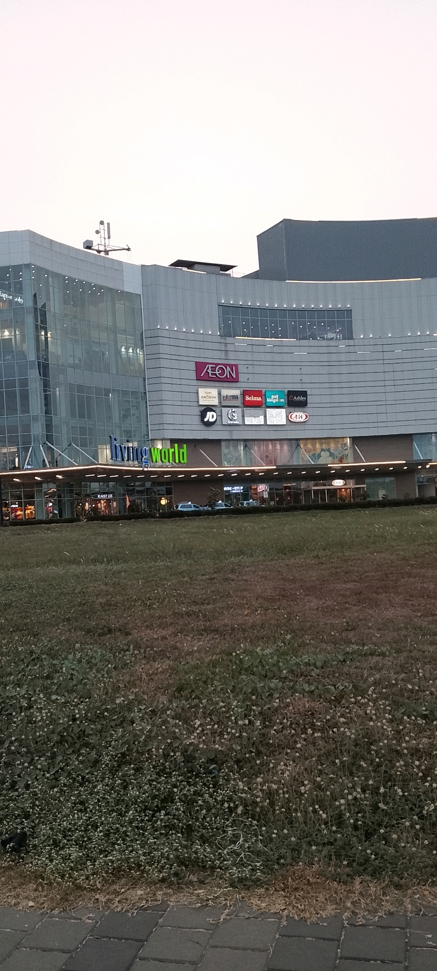 The image shows the exterior of a modern shopping mall identified by signage as "AEON" and "living world". The building features a glass facade on the left side and a grey, segmented facade on the right, adorned with multiple logos and signs for various shops including "TOYI KINGDOM", "JD", "Selma", "Ashley", and "AW". Below the main building, a covered walkway with a row of lights runs along the storefronts, which are mostly glass-fronted and appear to be shops or restaurants. Cars are visible parked in front of the building. In the foreground, a grassy area with sparse vegetation and some small white flowers stretches out, leading to a paved walkway made of dark rectangular tiles at the very bottom of the image. The sky above is overcast and pale. The scene is likely a commercial area in the city of Gunung Putri, Indonesia, given the "living world" branding which is known to be present there.