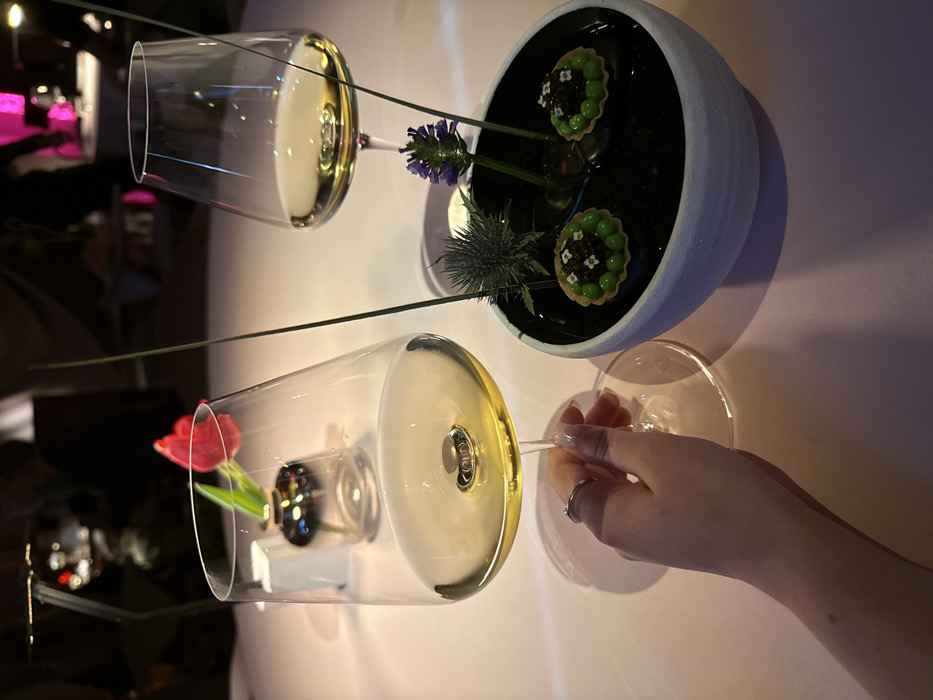 The image depicts an upscale dining or lounge setting in Dubai, United Arab Emirates, characterized by dim ambient lighting. A female's hand, adorned with a ring on the index finger, holds a clear stemmed wine glass filled with a golden-yellow liquid, likely white wine. A second identical wine glass containing the same liquid sits on the light-colored tabletop to the right. Between the two glasses, a decorative centerpiece rests in a white, textured bowl with a dark, possibly liquid-filled interior. This centerpiece features a spiky thistle, a purple lavender flower, two thin green stalks, and two small, round appetizers. These appetizers are green-filled tartlets, each topped with black caviar and small white edible florets. In the background, a small, reflective silver vase holds a single red tulip, positioned next to a minimalist, angular metallic table lamp. Blurred figures and magenta-hued lighting indicate other patrons and areas of the venue.