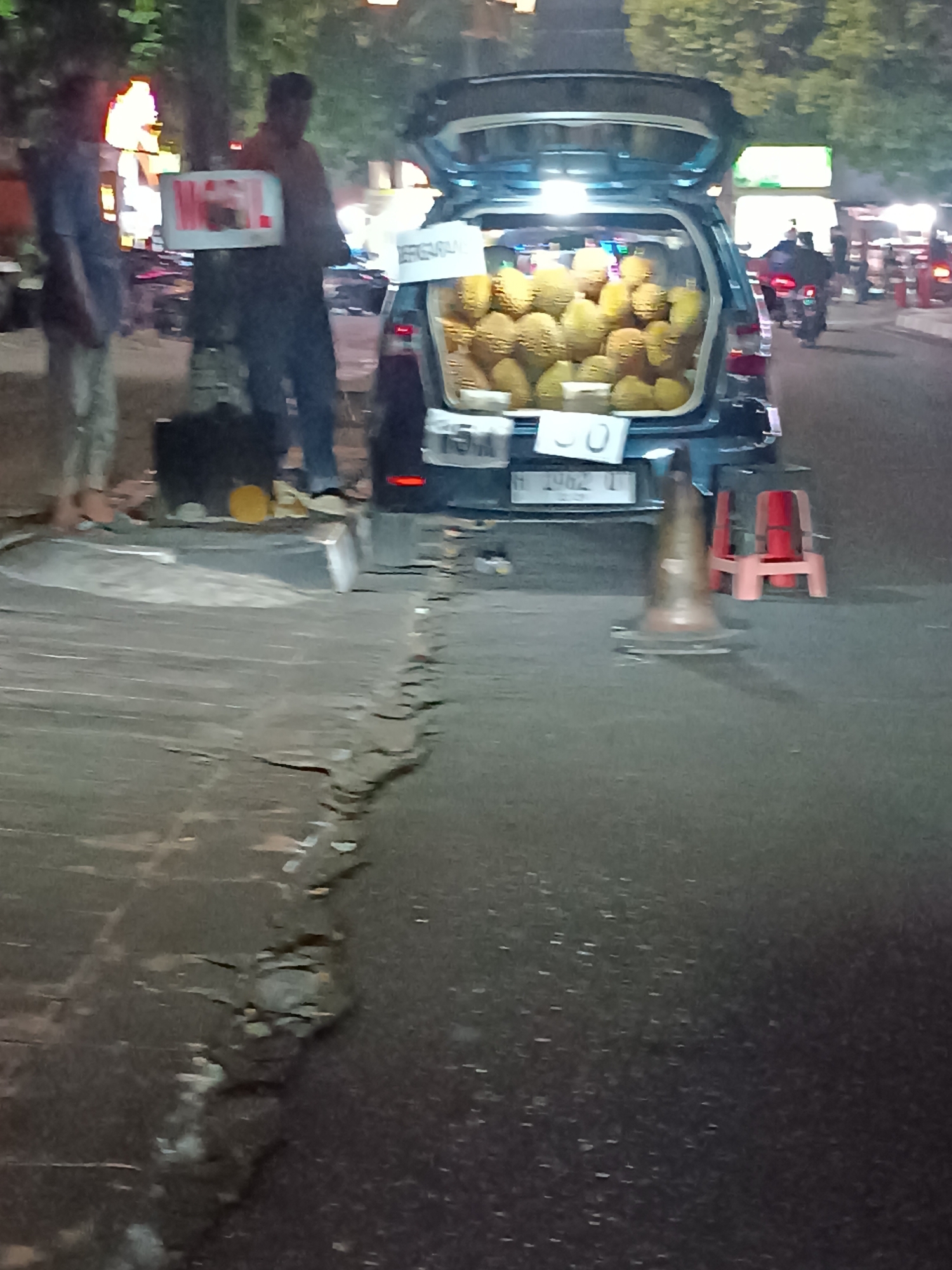 The image depicts a nighttime street scene in Yogyakarta, Indonesia, likely in an area where street vendors operate. In the foreground, the cracked asphalt of a road is visible. To the left, a person in a blue shirt and light-colored pants stands near a makeshift stall. The stall displays a sign with red and white lettering, featuring the word "MOL!". Behind this stall, another person in a dark shirt and pants is also present.
Dominating the center of the image is a dark-colored hatchback car with its trunk open, revealing a substantial quantity of durian fruit packed inside. A white sign with dark text, possibly indicating a price or vendor name, is affixed to the trunk lid. Two small, brightly colored red stools are positioned behind the car, along with a traffic cone. In the background, streetlights illuminate the scene, and the silhouettes of trees and other vehicles, including a motorcycle, can be seen. The overall impression is of a roadside durian vendor operating late at night.