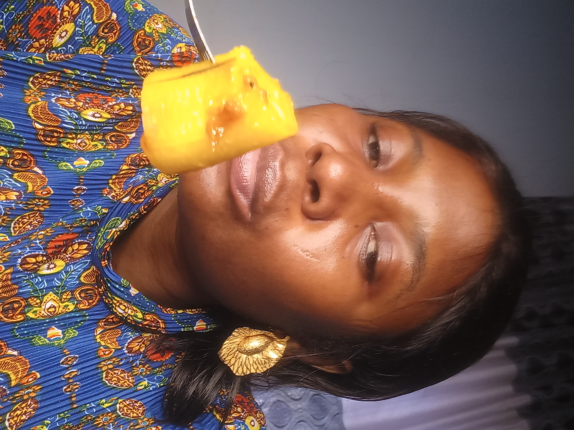 A medium shot captures a Black woman, with dark hair and brown eyes, in a close-up self-portrait. She is looking towards the right side of the frame with her mouth slightly open, as if about to eat a piece of yellow food impaled on a fork. She wears a large, gold, leaf-shaped earring on her right ear. Her blue, pleated dress is adorned with a vibrant pattern of red, yellow, and white floral and paisley designs. The background is divided into two sections: the left side is a solid dark gray, and the right side features a purple curtain with a textured pattern. The lighting appears to be natural, coming from the front and slightly to the left, illuminating her face and casting subtle shadows. The image is slightly grainy.