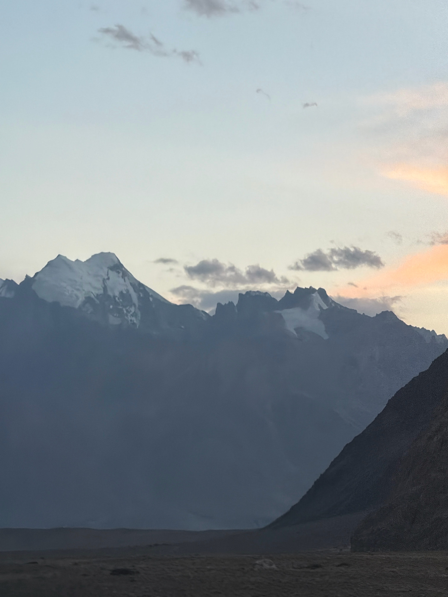 The image depicts a wide, panoramic view of a mountainous landscape at dusk. The sky is a gradient of soft blues and pale oranges, with a few wispy clouds. Dominating the background are jagged, snow-capped mountains shrouded in a dark, atmospheric haze. The peaks are sharply defined against the sky, with white snow contrasting with the dark rock. In the foreground, a flat, barren plain stretches towards the mountains. A steep, dark slope is visible on the right side of the frame, suggesting a foreground element of the landscape. The scene is devoid of any human presence or man-made structures, conveying a sense of natural, remote wilderness. The lighting suggests either sunrise or sunset, with the fading light casting a somber and majestic mood over the scene. The location is identified as Stongde, India, a remote region known for its stunning natural beauty and mountainous terrain.