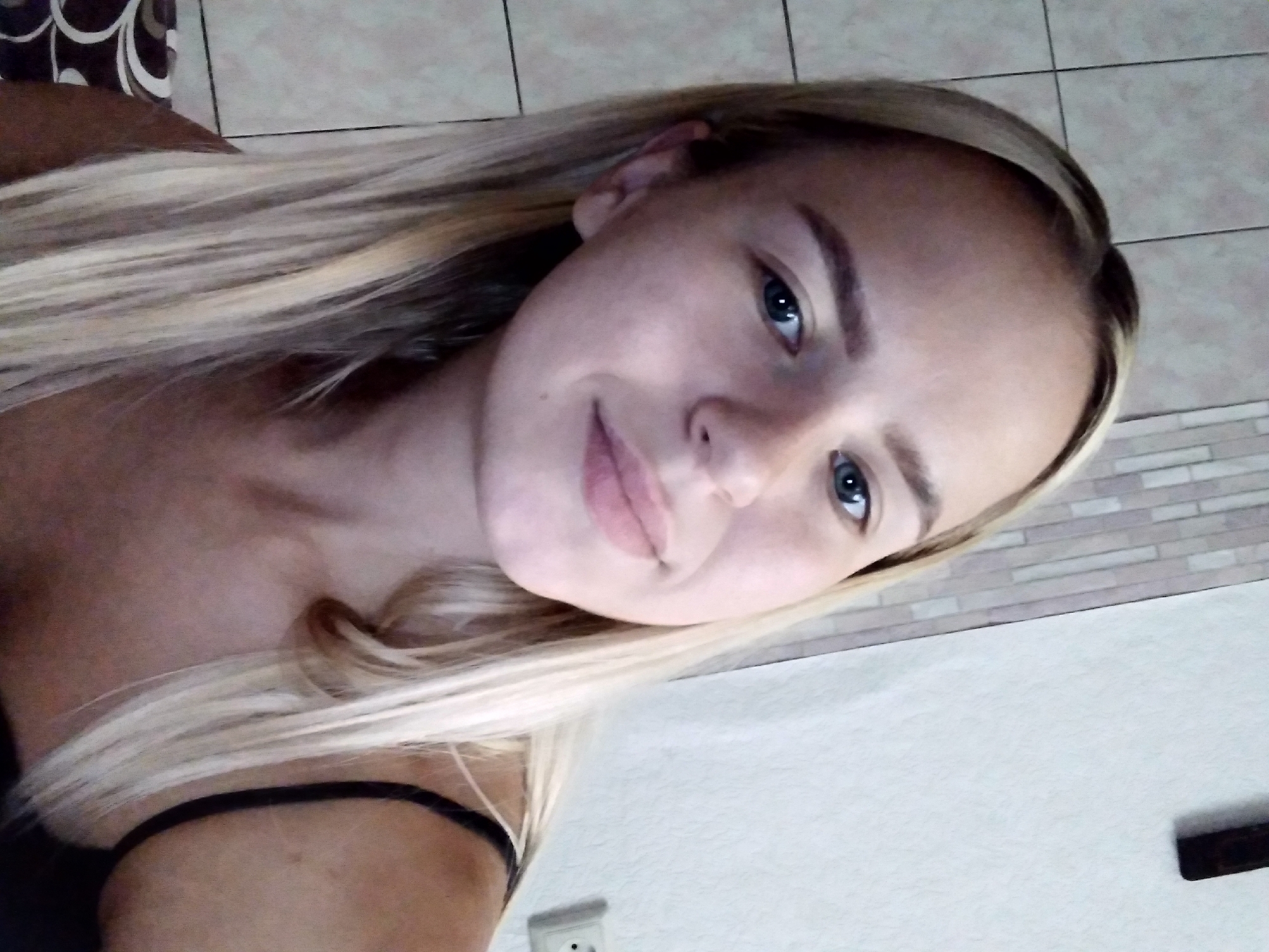 A close-up, head-and-shoulders selfie of a young Caucasian woman with blonde hair and blue eyes is presented. She is looking directly at the camera with a slight smile. The background includes a tiled wall on the left and a plain white wall on the right. A dark object, possibly a shelf or decorative item, is visible on the upper right portion of the white wall. A light-colored electrical outlet is also present on the white wall. The woman is wearing a black tank top. The image appears to have been taken indoors, possibly in a residential setting in Radisne, Ukraine.