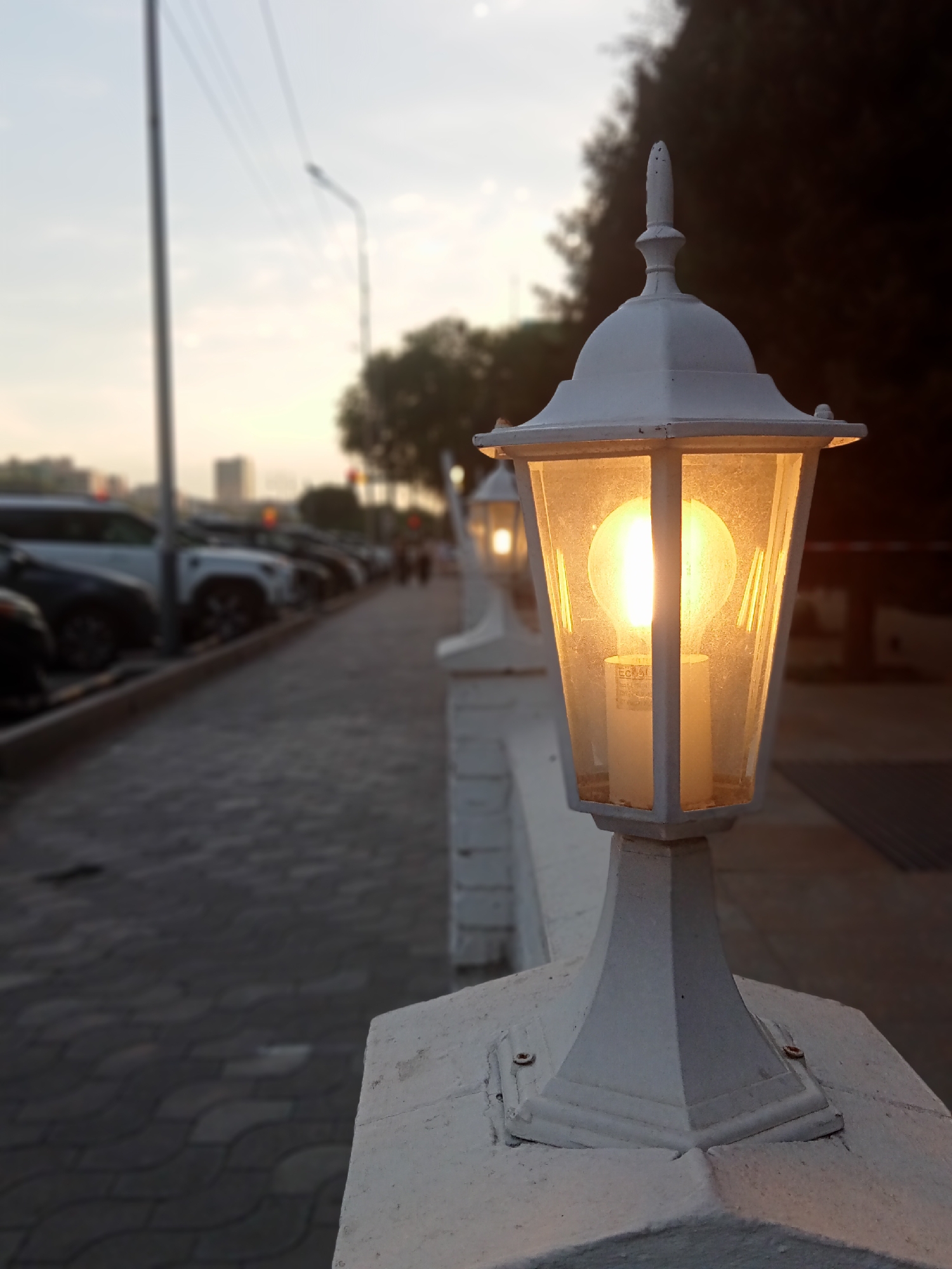 An outdoor scene, likely a walkway or pathway in Almaty, Kazakhstan, is captured at dusk or early morning. The foreground is dominated by a white, ornate lamppost with a glowing incandescent bulb inside its glass enclosure. The lamppost is mounted on a white, low concrete wall, with visible screws attaching it. Beyond the lamppost, the walkway is paved with interlocking grey concrete blocks. To the left of the walkway, a row of parked cars, including SUVs and sedans, are visible, suggesting a parking area adjacent to the pathway. The background is softly blurred, showing trees, a street light pole, and distant city buildings. The sky is a muted blend of grey and yellow, characteristic of twilight. Several other lampposts, similar in design to the one in the foreground, are spaced along the wall, also illuminated. The overall atmosphere is serene and slightly melancholic, with a focus on the artificial light of the lamppost against the fading natural light.