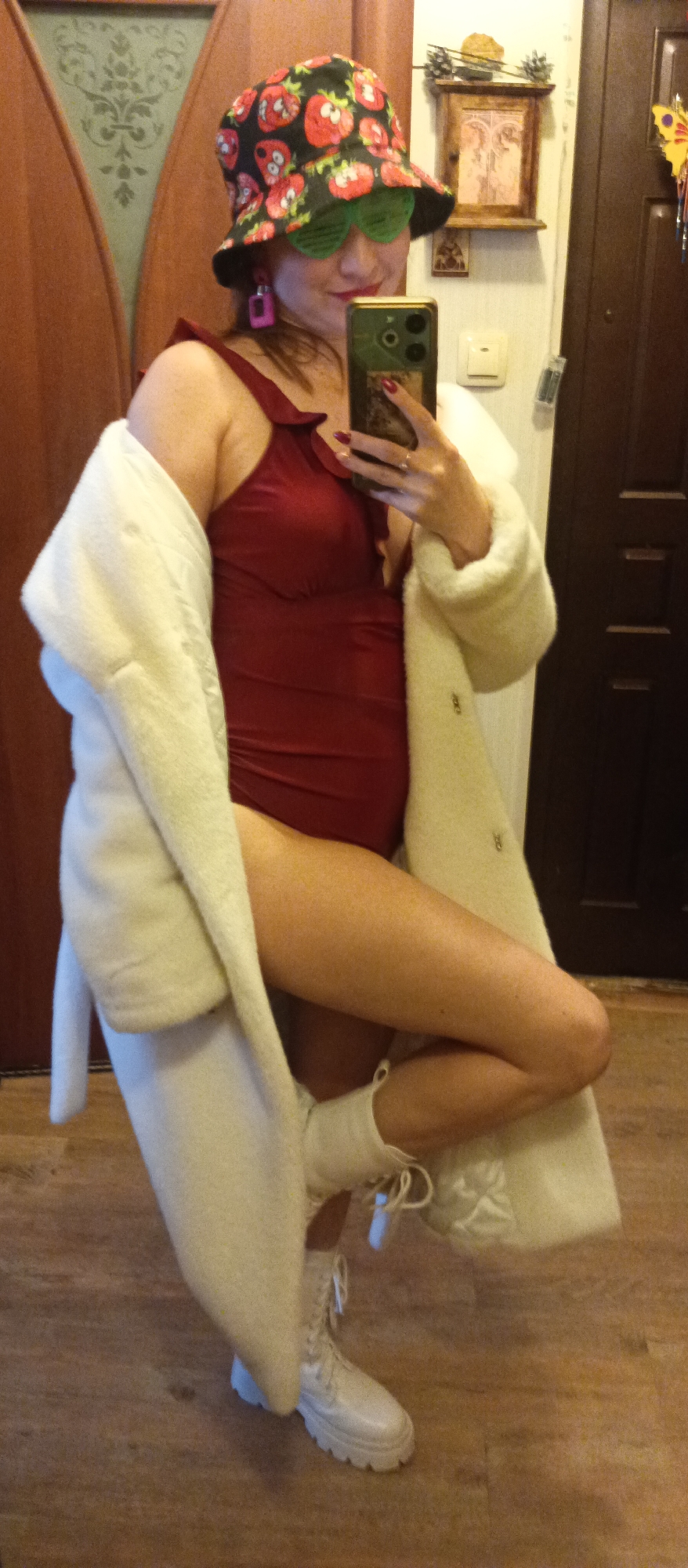 A person is posing indoors. They are wearing a dark red one-piece swimsuit, a large white coat draped over their shoulders, white combat boots, green sunglasses, a hat with a strawberry pattern, and pink earrings. The person is holding a smartphone up in front of them, presumably taking a selfie. The background shows a dark brown door and a wooden frame on the wall. The location is Unknown, Unknown.