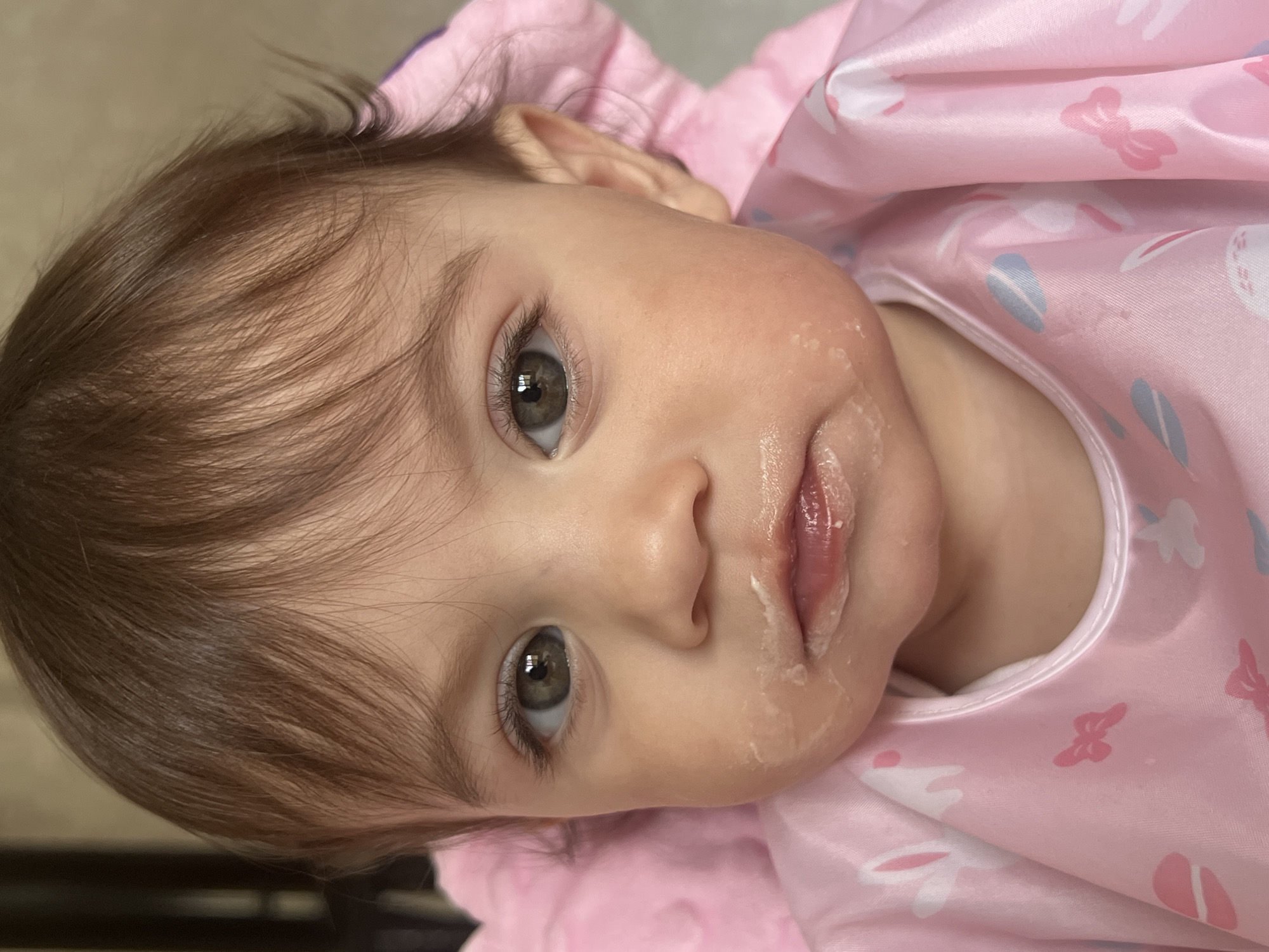 This close-up portrait captures a baby or young toddler in an indoor setting, potentially in Elena, Bulgaria. The baby has light brown hair and distinctive hazel or gray-green eyes, looking directly at the camera with a neutral, attentive expression. White, creamy food residue is visible around the baby's mouth and on the left cheek, suggesting a recently completed meal or snack. The baby is wearing a glossy, satin-like pink bib or smock adorned with patterns of white bunnies, blue oval shapes, and pink bows. The background is softly blurred, indicating an indoor environment. No text is visible in the image. The baby appears to be in a reclined or lying-down position.