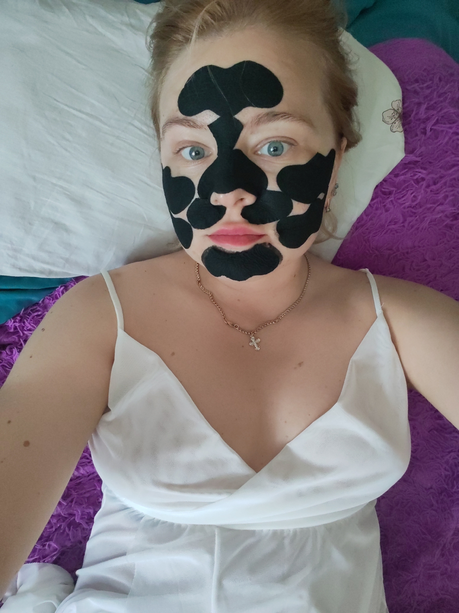 A fair-skinned woman with light blue eyes and blonde hair is lying down, looking up at the camera. Her face is partially covered with irregularly shaped black patches, suggestive of a face mask treatment. She is wearing a white, thin-strapped top or dress and a delicate gold necklace with a cross pendant. She is lying on a bed with a white pillow and a textured purple blanket. The background also includes a turquoise-colored object. There is no information given to imply this image takes place in Izhevsk, Russia. The provided context offers no details related to the city or its location.