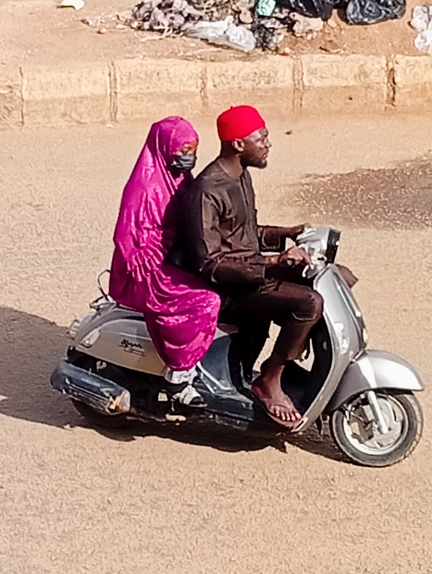 In Bukuru, Nigeria, a man wearing a red cap and a dark brown tunic rides a silver scooter. He is looking to his right. Behind him, a woman in a bright pink headscarf and dark face mask is seated on the scooter. She is wearing a matching bright pink garment. The scooter is traveling on a dusty, light brown road. In the background, there is a curb with some debris and trash piled on top.