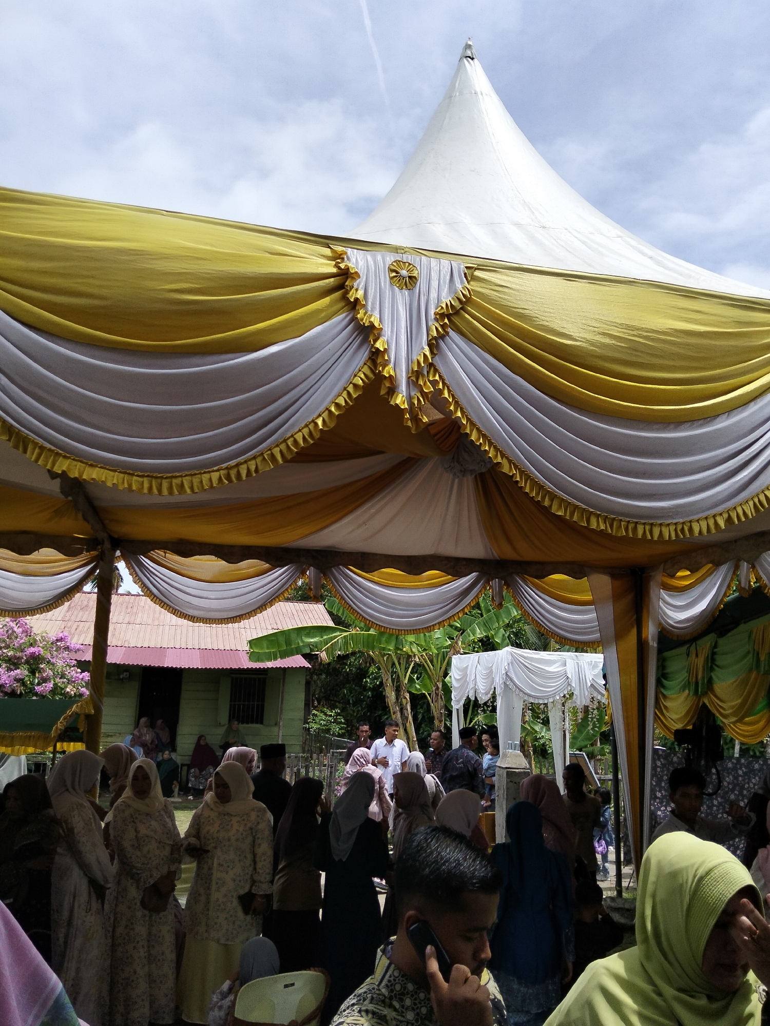 The image depicts an outdoor event, likely a gathering or celebration, taking place under a large white and yellow tent. The tent is adorned with decorative draping and ruffles. In the background, a modest green building with a reddish-brown roof is visible, suggesting a rural or suburban setting in Cottrueng, Indonesia. Several people are present, many of them women wearing headscarves and traditional attire. Some individuals are seen standing, while others appear to be seated or mingling. The overall atmosphere seems to be that of a communal event, possibly a wedding or cultural festival, given the festive decorations and the gathering of people. The sky above is partly cloudy, indicating daytime.