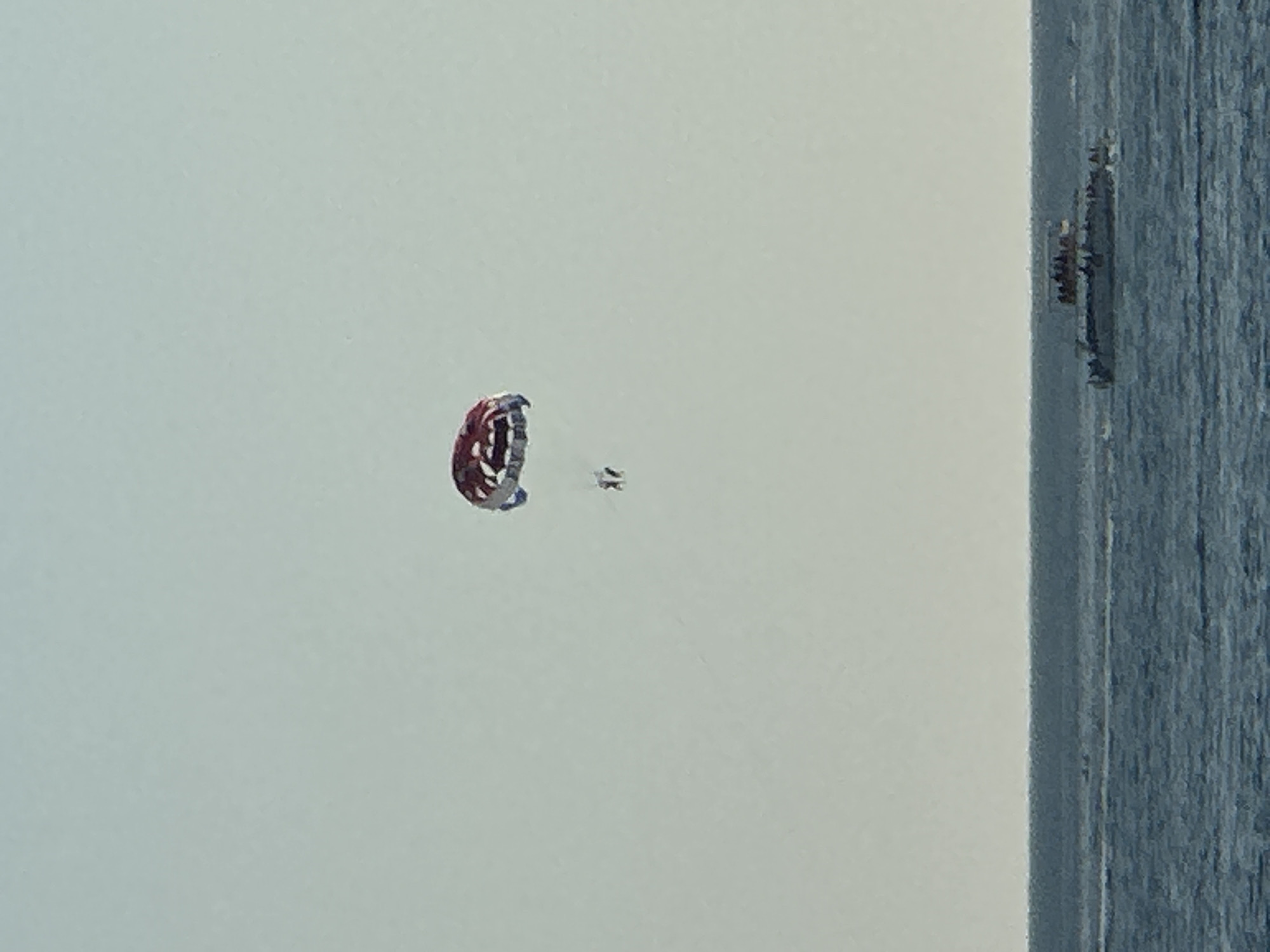 An outdoor, wide shot shows a parasail drifting in the sky above the Adriatic Sea. The parasail, which is red and white, has two people riding below it. A catamaran, with several people visible on its deck, sails on the water. The scene appears to be in Budva, Montenegro, given the geographical context. The water is choppy and blue, with a thin line of the horizon separating it from the pale sky.