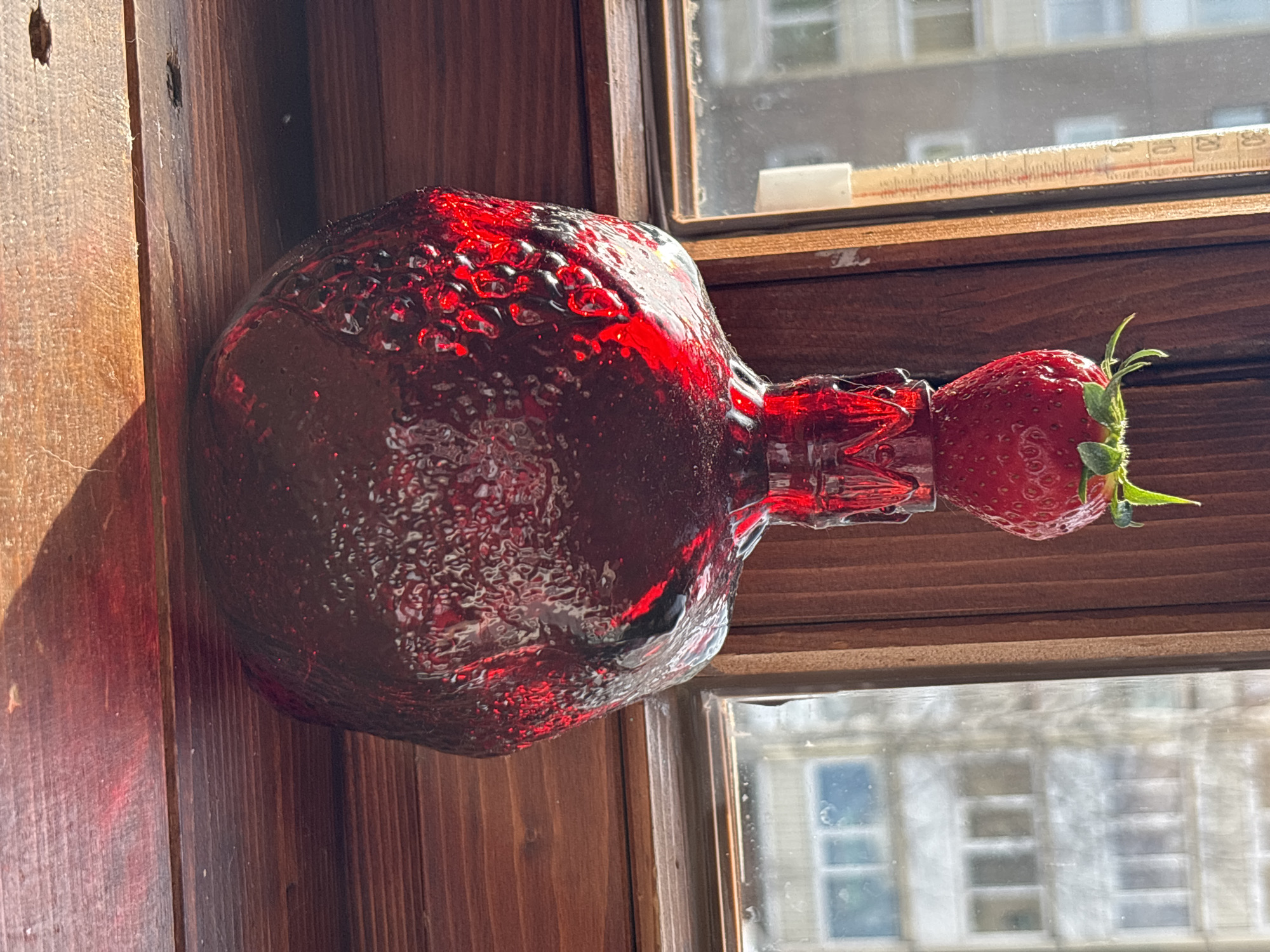 A red glass bottle is positioned horizontally on a wooden windowsill. The bottle has a bulbous shape and a narrow neck. A strawberry is attached to the bottle's neck. Outside the window, a multi-story building is visible. The windowsill is situated within a room with wooden walls. The location is Марковский, Russia.