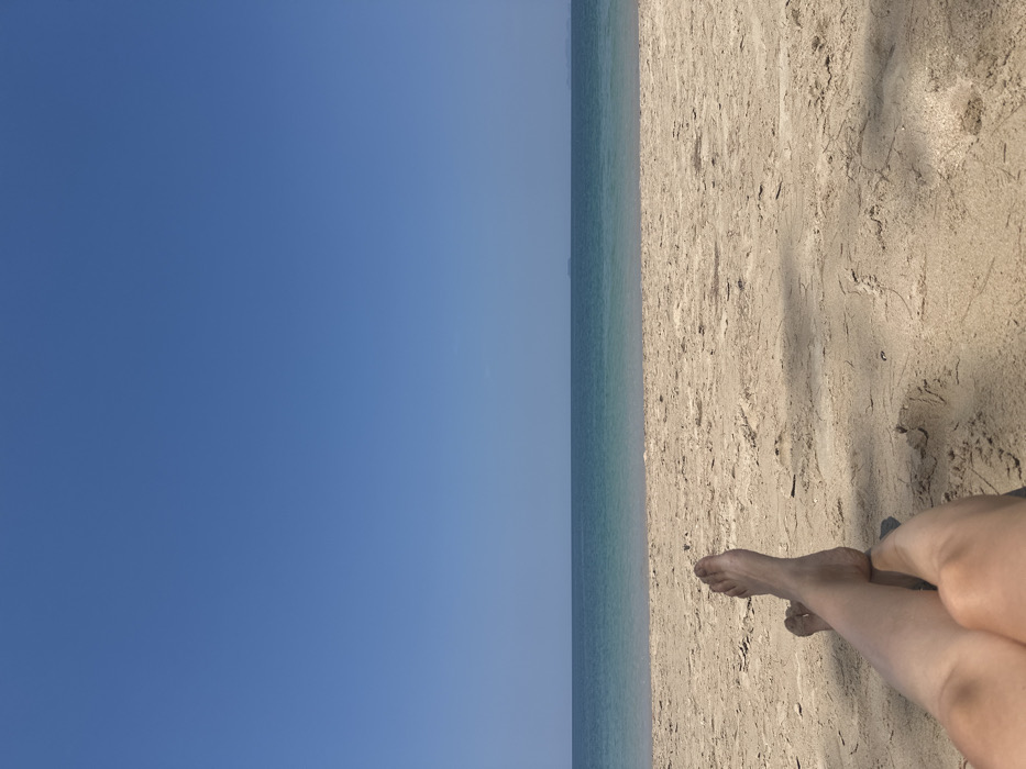 The scene depicts a daytime view from a sandy beach in Phra Ae, Thailand. In the immediate foreground, the bare lower legs and feet of an individual are visible, resting on light-colored sand. The legs are crossed at the ankles, and shadows from an unseen overhead object are cast across the sand. The midground comprises a wide expanse of light brown beach, transitioning into the ocean. The water appears in a gradient, from a lighter turquoise near the shore to a deeper blue further out. The background is dominated by a clear, cloudless bright blue sky, with a distinct horizon line. Two faint, distant landmasses or islands are barely discernible on the horizon against the sky.