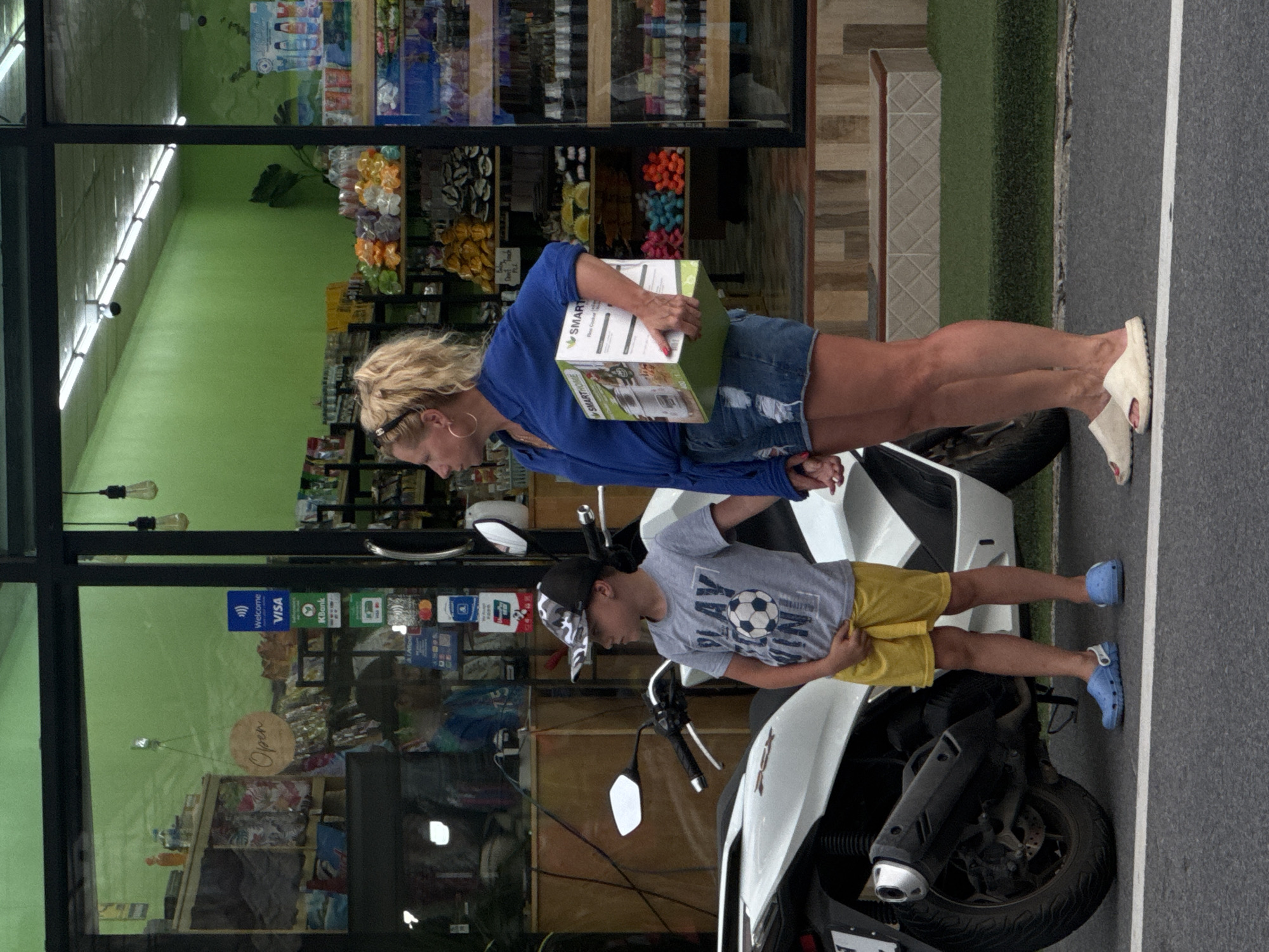 A woman and a young boy are standing next to a white motorcycle in front of a store. The woman is wearing a blue shirt and denim shorts, holding a rectangular box. The boy is wearing a grey t-shirt, yellow shorts, and a camouflage baseball cap. They appear to be holding hands. The store has a green exterior and displays various products. Payment method signs, including Visa, are visible on the window. The ground is asphalt with a white line. The scene is located on a street in Karon, Thailand, suggested by the "Open" sign in Thai script.