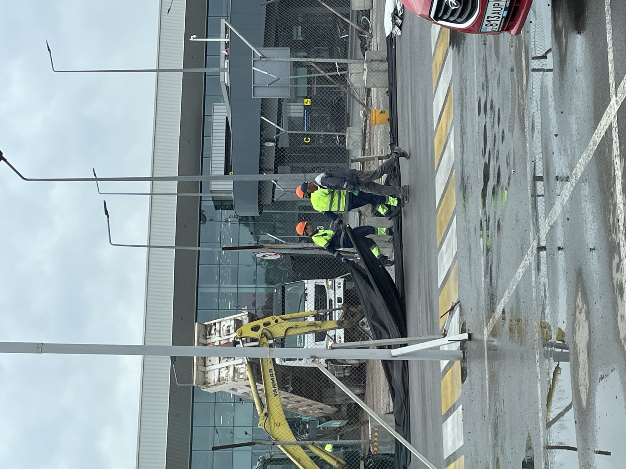 The image depicts a construction scene at an outdoor location, possibly near an airport due to the large modern building with glass facades in the background. The ground is wet, indicating recent rain, with visible puddles and reflections. A red car with a license plate reading "813 AUP" is partially visible on the right side of the frame.
In the center of the image, three construction workers are present. Two of them, wearing orange hard hats and high-visibility vests over their work clothes, are handling a large piece of black tarp. One worker on the left is bent over, appearing to assist in dragging the tarp, while the worker in the middle, also in a high-visibility vest, is walking while holding onto the tarp. A third worker, wearing a cap and a grey jacket, is walking away from the other two.
Behind the workers, a yellow excavator with the brand "YANMAR" visible on its arm is positioned next to a white dump truck. The dump truck's bed is raised, and it appears to be in the process of unloading material. A chain-link fence runs across the mid-ground, separating the construction area from the foreground. Several concrete barriers and poles are also part of the construction site infrastructure.
The building in the background is a modern structure with a large expanse of glass windows. Streetlights are visible in the background, as are other fencing elements and possibly more construction equipment. The overall scene suggests ongoing construction or maintenance work in an urban or semi-urban setting.
While the image provides visual clues, the specific location in Almaty, Kazakhstan cannot be definitively determined from the visual information alone. However, the presence of the license plate "813 AUP" could potentially be used to trace the vehicle's registration and, by extension, its origin, if such information were publicly accessible and linked to Almaty.