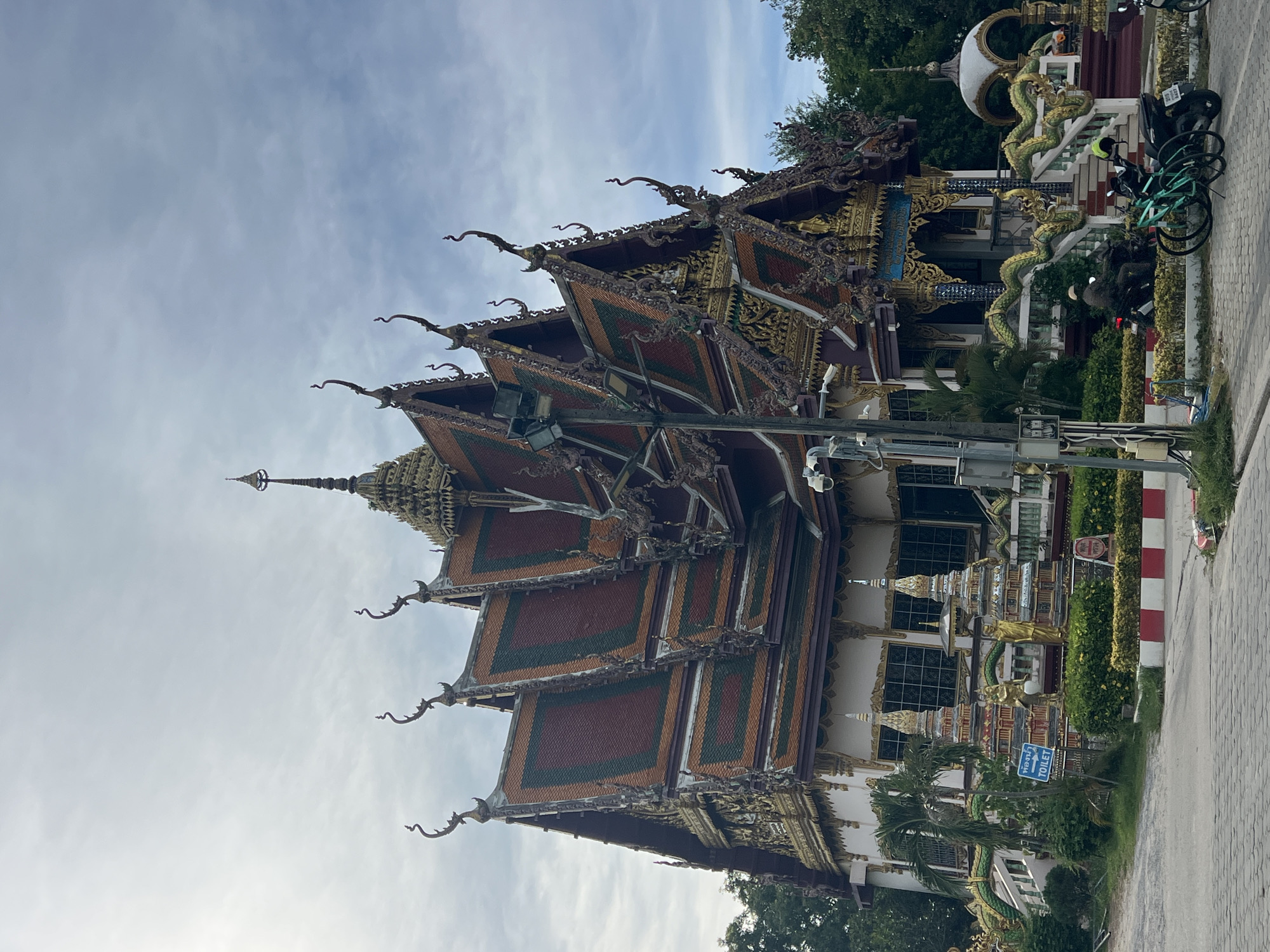 This is a photograph of the Wat Plai Laem temple located in Baan Plai Laem, Thailand. The temple is characterized by its ornate, multi-tiered roof with vibrant orange, green, and brown tiles, adorned with decorative finials and sculptures. The facade of the temple is predominantly white with intricate gold detailing. Two golden Buddha statues stand on the temple grounds near the entrance, flanked by decorative structures. A prominent sign with Thai script and the English word "TOILET" is visible to the left of the temple. The temple is surrounded by lush green trees and foliage. In the foreground, a paved area shows a street with a motorcyclist in motion and two parked bicycles. A lamppost with security cameras is positioned in front of the temple. The sky above is overcast with scattered clouds.