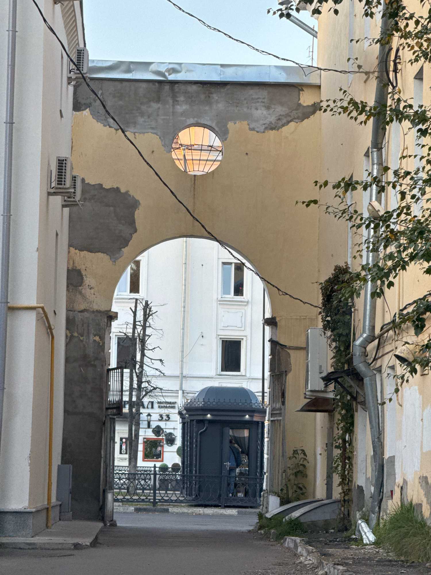 The image shows an archway in a city, likely Tver, Russia, leading to a courtyard. The archway is made of weathered yellow stucco, with patches of exposed concrete. Above the arch, a circular window with a stained-glass design is visible, illuminated from behind. On the left side of the arch, there are two air conditioning units mounted on a cream-colored building. A black wire runs diagonally across the scene.
Through the arch, a white building with multiple windows can be seen. In front of this building, there is a small, dark structure resembling a kiosk or guardhouse, with decorative metalwork. A black wrought-iron fence with a Greek key pattern separates the walkway from the courtyard. A bare tree stands behind the fence.
To the right of the arch, a yellow building with peeling paint is visible. A metal downspout runs vertically down the side of this building, with ivy growing on it. Another air conditioning unit is mounted higher up on this building. The ground in the foreground is asphalt with a cracked concrete curb. The overall impression is one of an older urban environment, possibly showing signs of neglect and renovation. There are no people clearly depicted in the image, although figures are partially visible through the windows of the kiosk. The sky is overcast.