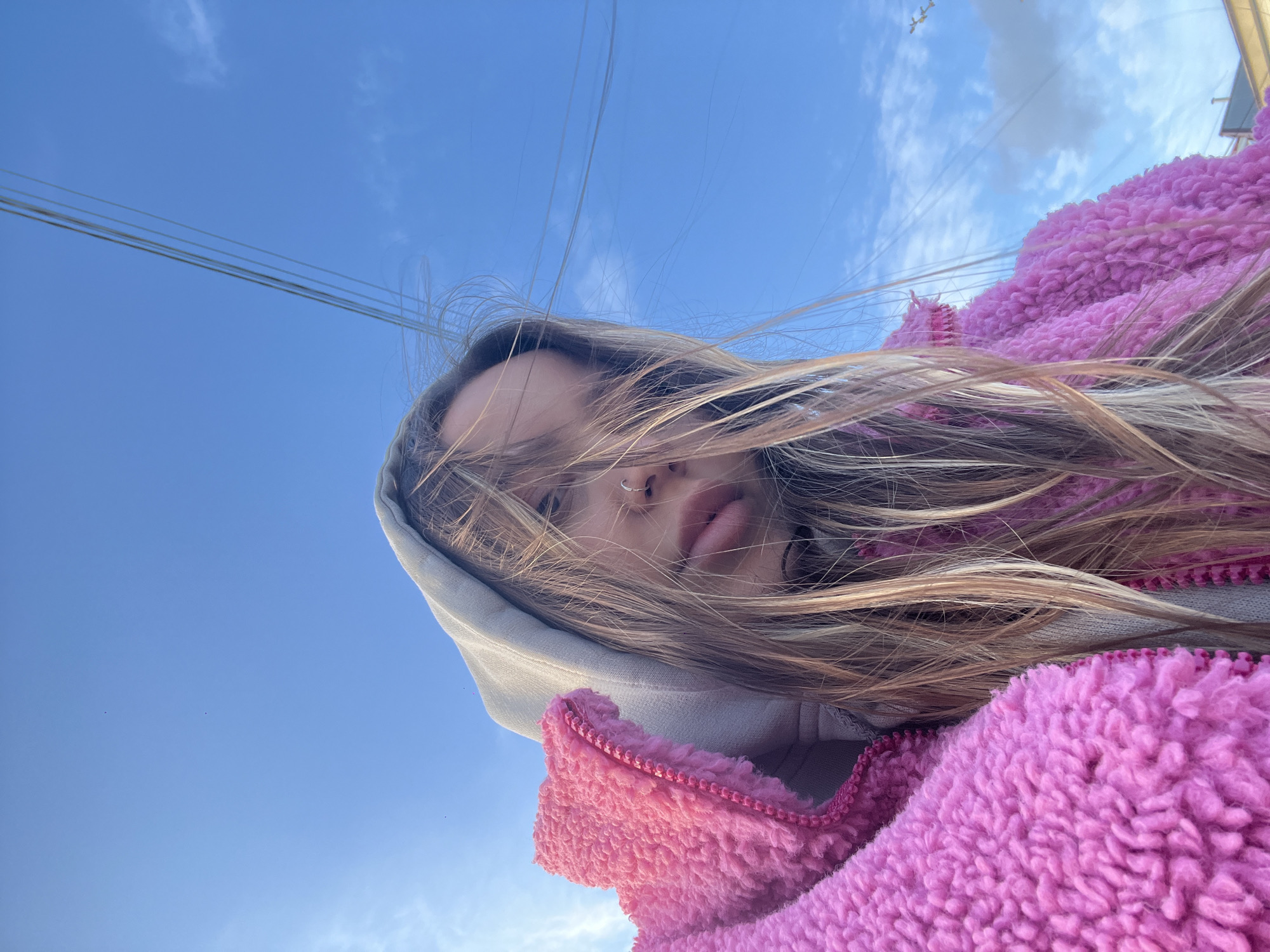 A low-angle upward selfie shot shows a young woman wearing a light gray hoodie and a pink, fuzzy fleece jacket. Her blonde hair is partially obscuring her face. A nose ring is visible. In the background is a clear blue sky with thin white clouds and power lines. The location, Novopokrovskaya, Russia, is not discernable from the image.