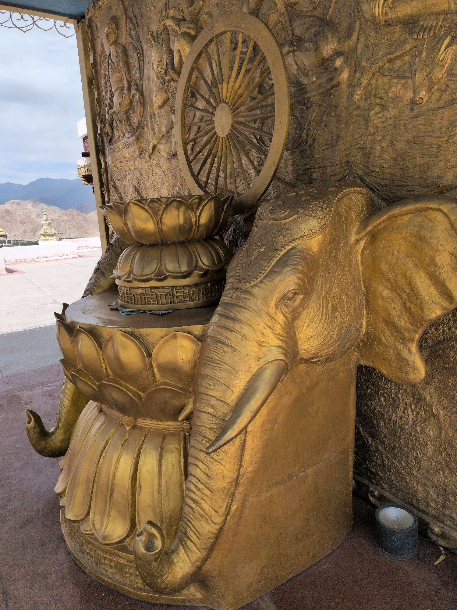 This is a close-up, eye-level, medium shot of a golden, intricately carved sculpture in Martselang, India. The sculpture features a prominent elephant head on the right, facing left, with its trunk extending downwards. To the left of the elephant head is a tiered lotus flower design, which serves as a base. Behind the lotus design, on the wall of the sculpture, is a bas-relief carving that includes a Dharma wheel, a Buddhist symbol, and depictions of human figures. In the background, a hazy mountain range is visible under a cloudy sky, with a stupa structure on the left. The ground is paved with reddish-brown tiles, and a small black floodlight is visible in the lower right corner.