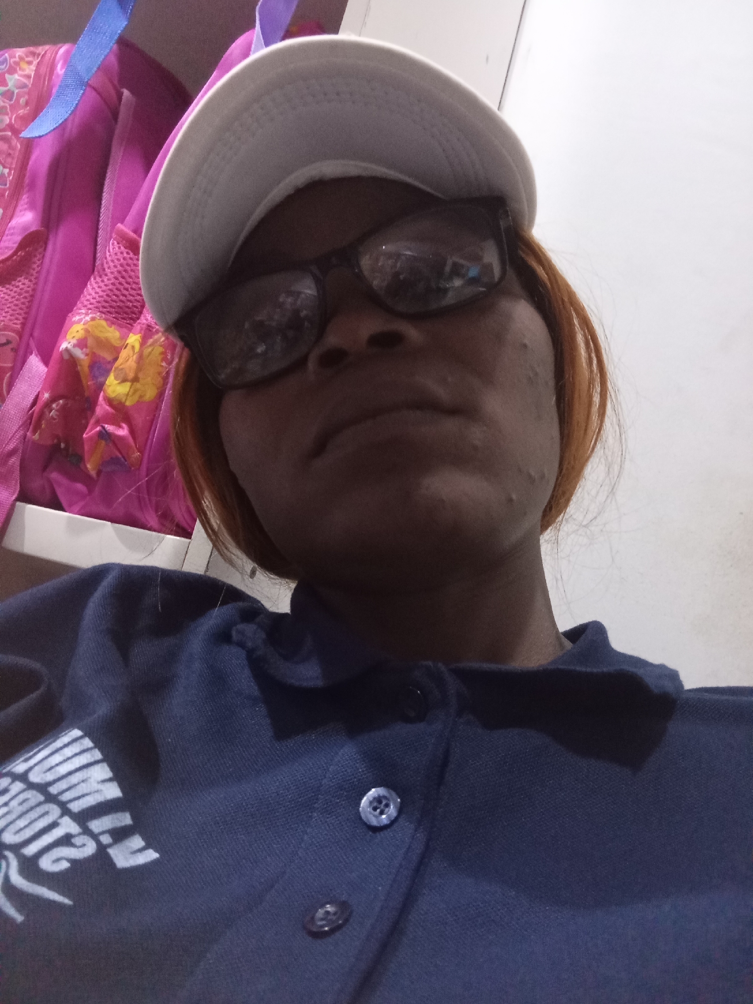 A young African woman is shown in a close-up, selfie-style photograph. She is wearing a white baseball cap and dark-rimmed glasses. Her auburn hair is visible around her face. She is wearing a dark blue polo shirt with a white graphic design on the left side of the chest. The graphic appears to be text and possibly a logo. Two buttons are visible on the placket of the shirt. In the background, a pink backpack with floral patterns and blue straps is partially visible. The setting appears to be indoors, with a plain white wall behind the woman. The image has a slightly grainy quality. The location is the city of Hawayi, Nigeria.