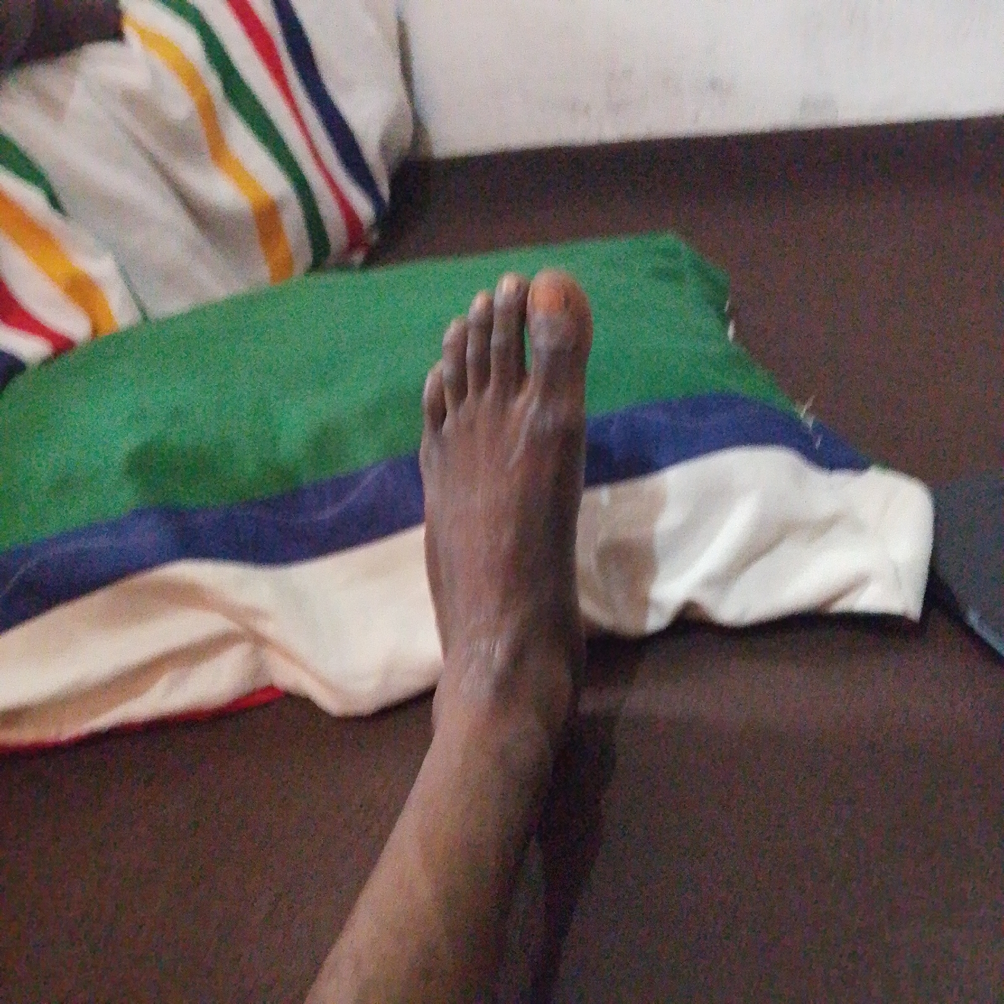 A close-up, overhead shot shows a person's left foot and lower leg, which is dark-skinned, resting on a pillow. The pillow has a white base with horizontal stripes of green, blue, and red. The pillow is lying on a dark brown surface, possibly a bed or sofa. The lighting appears dim and the image is slightly out of focus. This image was taken in Jalingo, Nigeria.