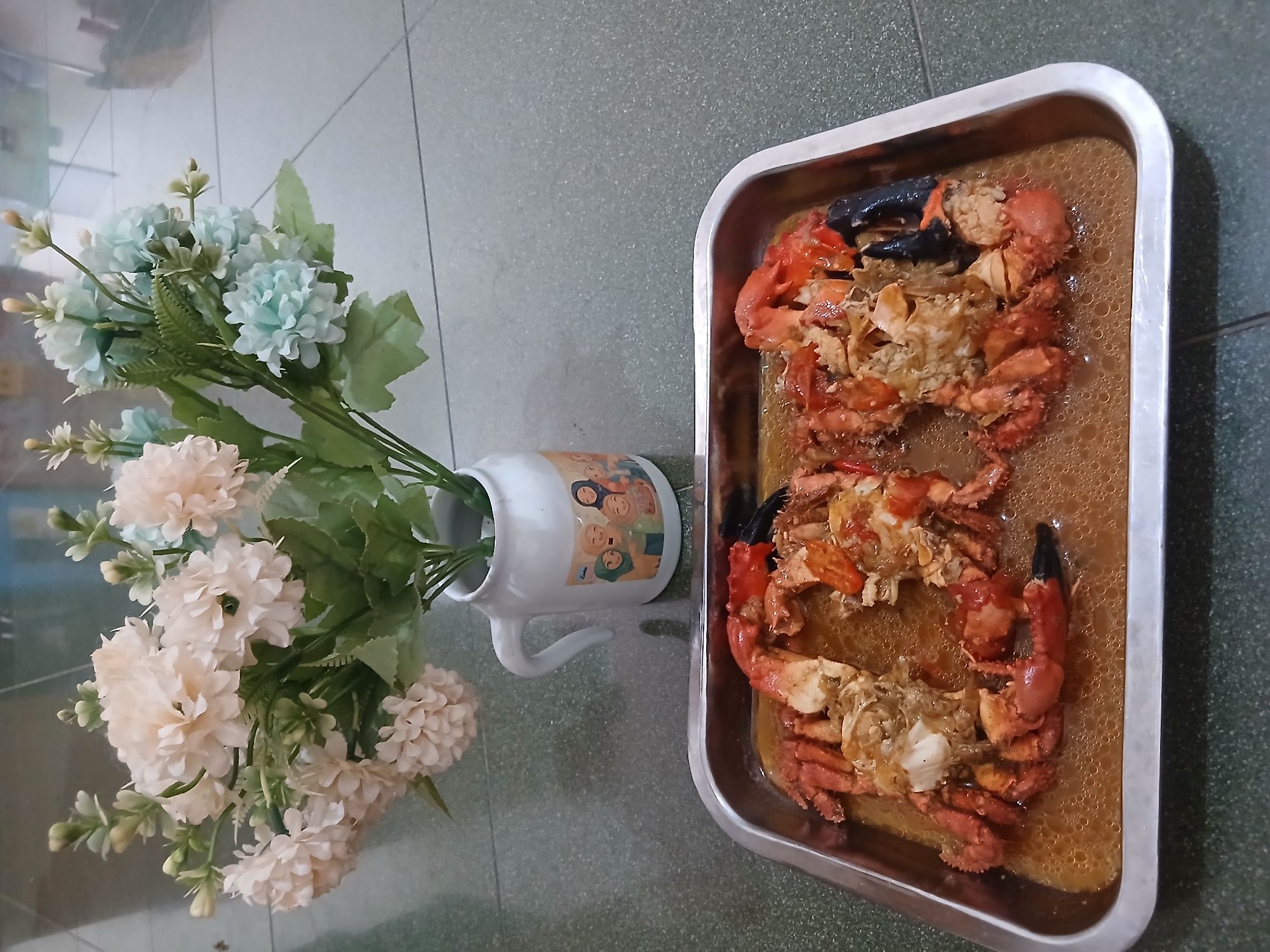 The image displays two cooked crabs, broken into pieces and presented in a metal tray filled with a savory brown sauce. The crabs are a vibrant orange-red color, with visible claws and segmented bodies. The sauce appears to have small bubbles on its surface, suggesting it is warm. Behind the tray, a bouquet of artificial flowers is arranged in a white ceramic pitcher adorned with illustrations of people wearing hijabs. The pitcher is positioned to the right of the flowers. The setting appears to be an indoor space with a tiled floor, possibly in a residential or restaurant environment in West Jakarta, Indonesia, given the context of the flower design. The overall lighting is soft and diffused, casting a gentle glow on the food and floral arrangement.