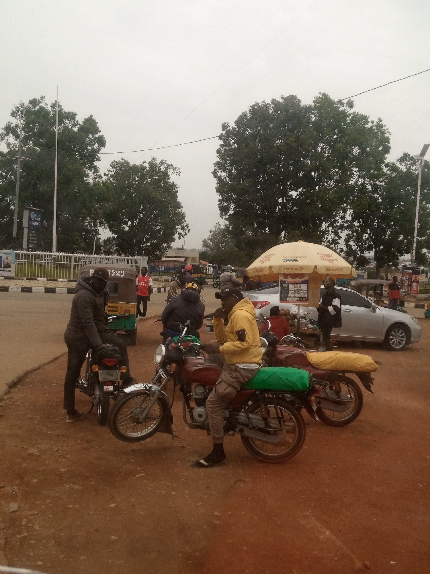 In this image, several people are seen gathered around motorcycles and a car on a dusty road. The scene appears to be an outdoor market or transportation hub. In the foreground, two men are seated on motorcycles. One is wearing a yellow jacket and khaki pants, while the other, partially obscured, is in a dark jacket and helmet. Both motorcycles have bags strapped to the back.
Behind the motorcycles, a silver car is parked. To the left of the car, a man in a black and white jacket stands near a stall with a large yellow umbrella advertising "Lucozade." The stall displays various services, including "CABLE BILLS," "AIRTIME RECHARGE," and "DATA SUBSCRIPTION." Further in the background, other individuals and a tricycle are visible.
To the left of the frame, a building with signage that includes "PLACE CINEMAS" and "CINEMA" suggests a commercial area. The sky is overcast, indicating potentially cloudy or hazy weather. The overall impression is a busy street scene with vendors and people waiting or preparing for travel. The presence of multiple motorcycles suggests a mode of transportation common in the area. The visible text provides details about available services and businesses.