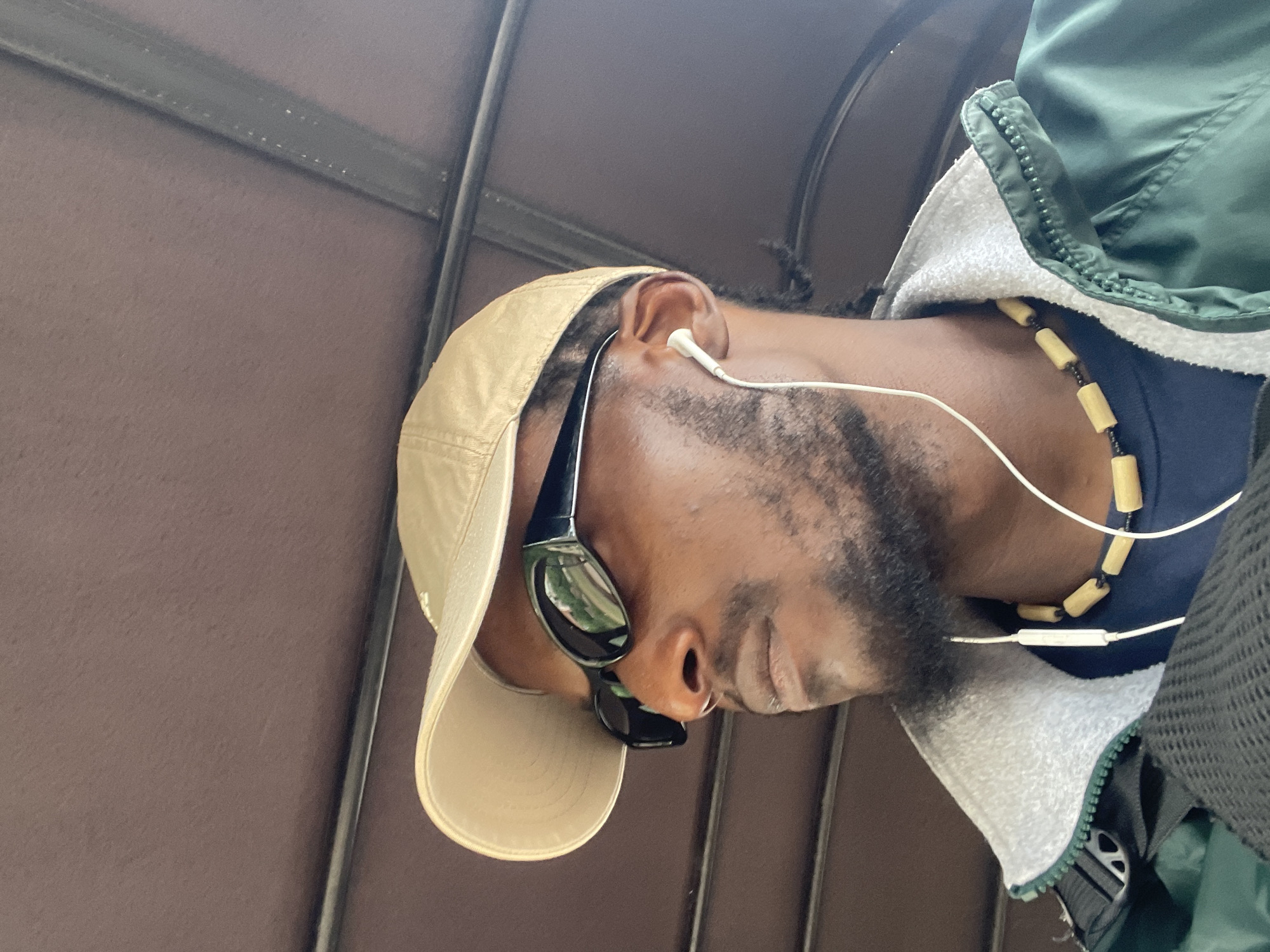 The image shows a close-up, low-angle shot of a Black man from the chest up. He is wearing a tan baseball cap, dark sunglasses with green reflections, and a green jacket over a dark blue shirt. A white-wired earbud is in his right ear, and a beaded necklace with light-colored cylindrical beads and black spacers is around his neck. The background appears to be the interior of a vehicle, with a dark brown padded surface and metal bars suggesting seating or a frame. The lighting suggests it might be daytime, and the mood is casual and relaxed. There is no visible text in the image. The location context is Jos, Nigeria.