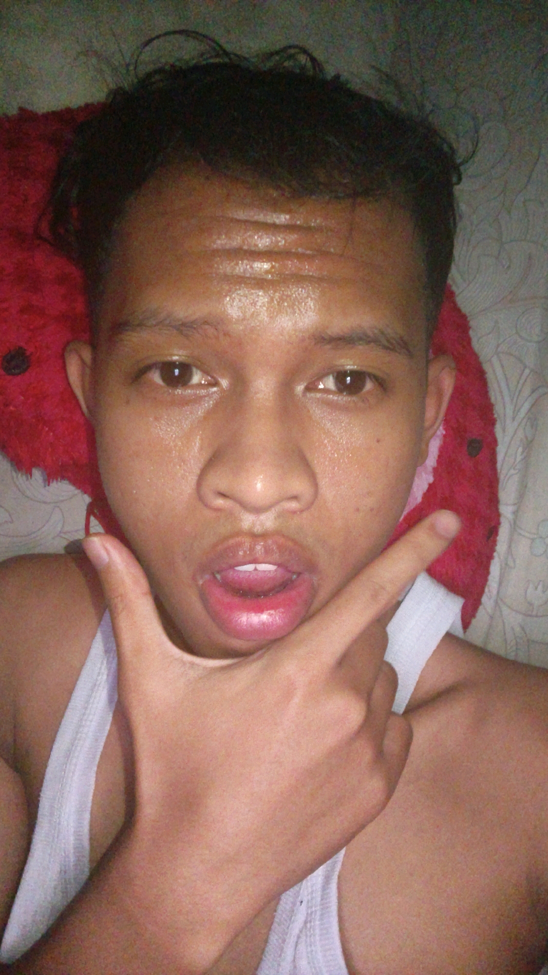 A young person is lying on their back, possibly in bed, with a red pillow behind their head. They are wearing a white tank top. Their face is sweaty, and they are looking directly at the camera with their mouth slightly open, revealing teeth. Their right hand is raised, with the index finger pointing towards their mouth. The background appears to be a patterned sheet or wall. The lighting is artificial and casts shadows on the person's face. The overall impression is a casual, candid selfie taken in an indoor environment. This image was taken in Pundungsari, Indonesia.