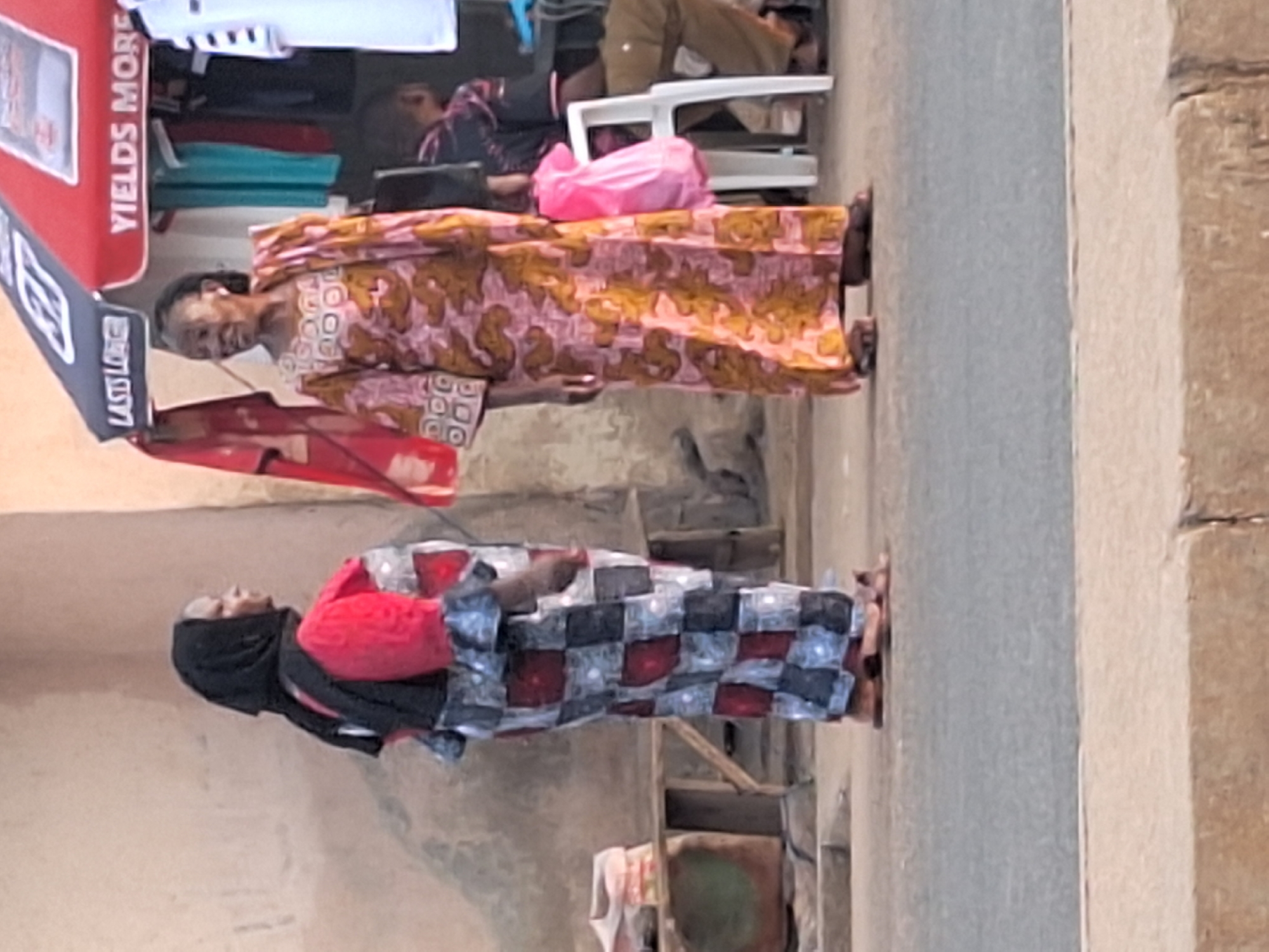 In Sabon Tasha, Nigeria, two women are standing outside, one wearing a pink and gold patterned dress, the other in a black headscarf and a checkered dress with red accents. A red and white vending machine stands nearby. In the background, people are sitting. The environment appears to be a street or public area.