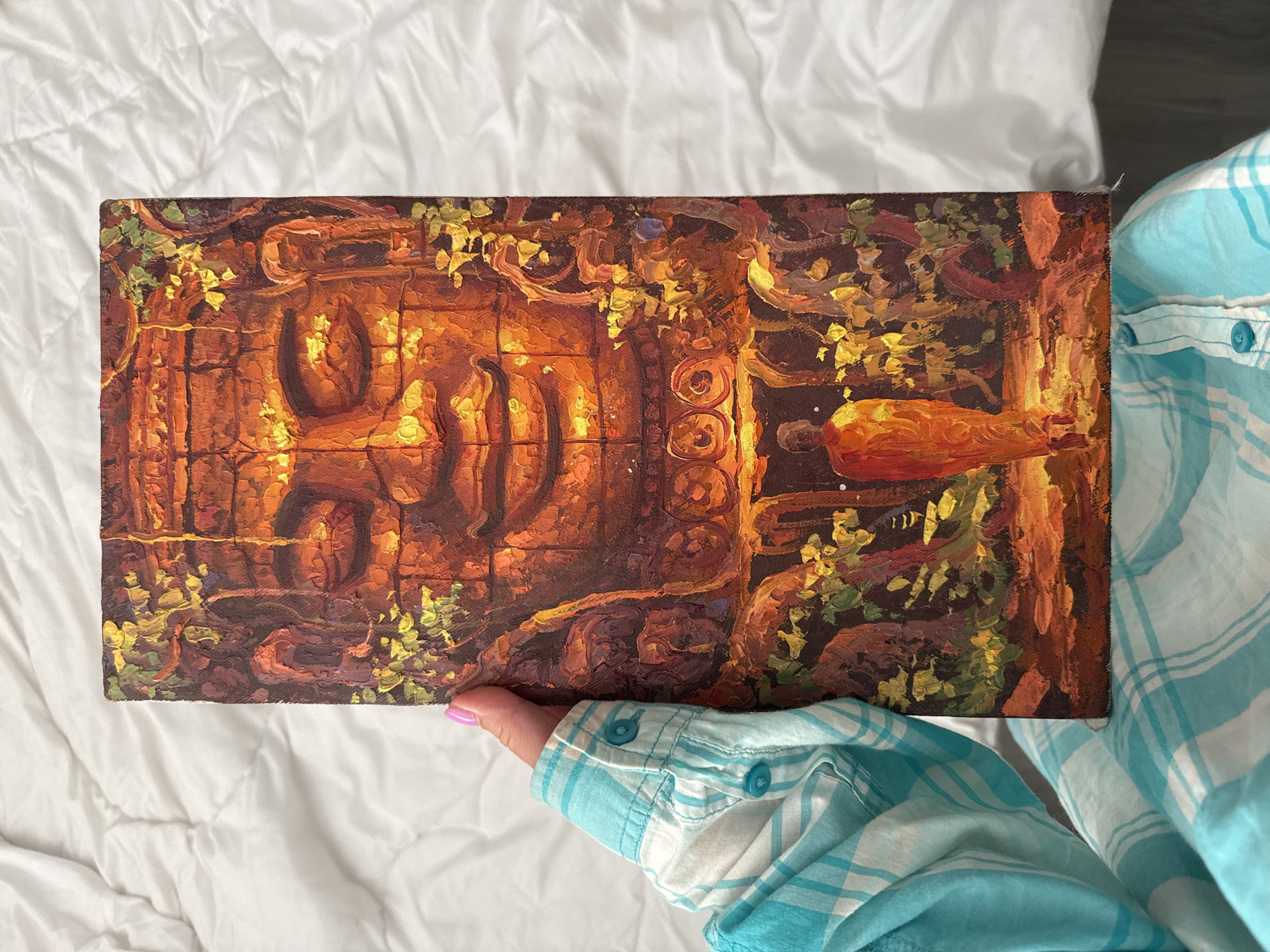 A person is holding a rectangular painting of a large stone face, possibly of a Buddha, overgrown with vegetation. The face is the dominant feature, rendered in warm, earthy tones of orange and brown, with a serene expression. Below the face, a small figure of a person in an orange robe walks towards it. The painting is textured, suggesting it is oil on canvas. The background is a white, quilted surface, likely a bed. The person holding the painting is wearing a blue and white plaid shirt. The image is cropped at the top and right, suggesting the painting is being presented for viewing. The location in the city of Khan Mean Chey, Cambodia, is not discernible from the image.