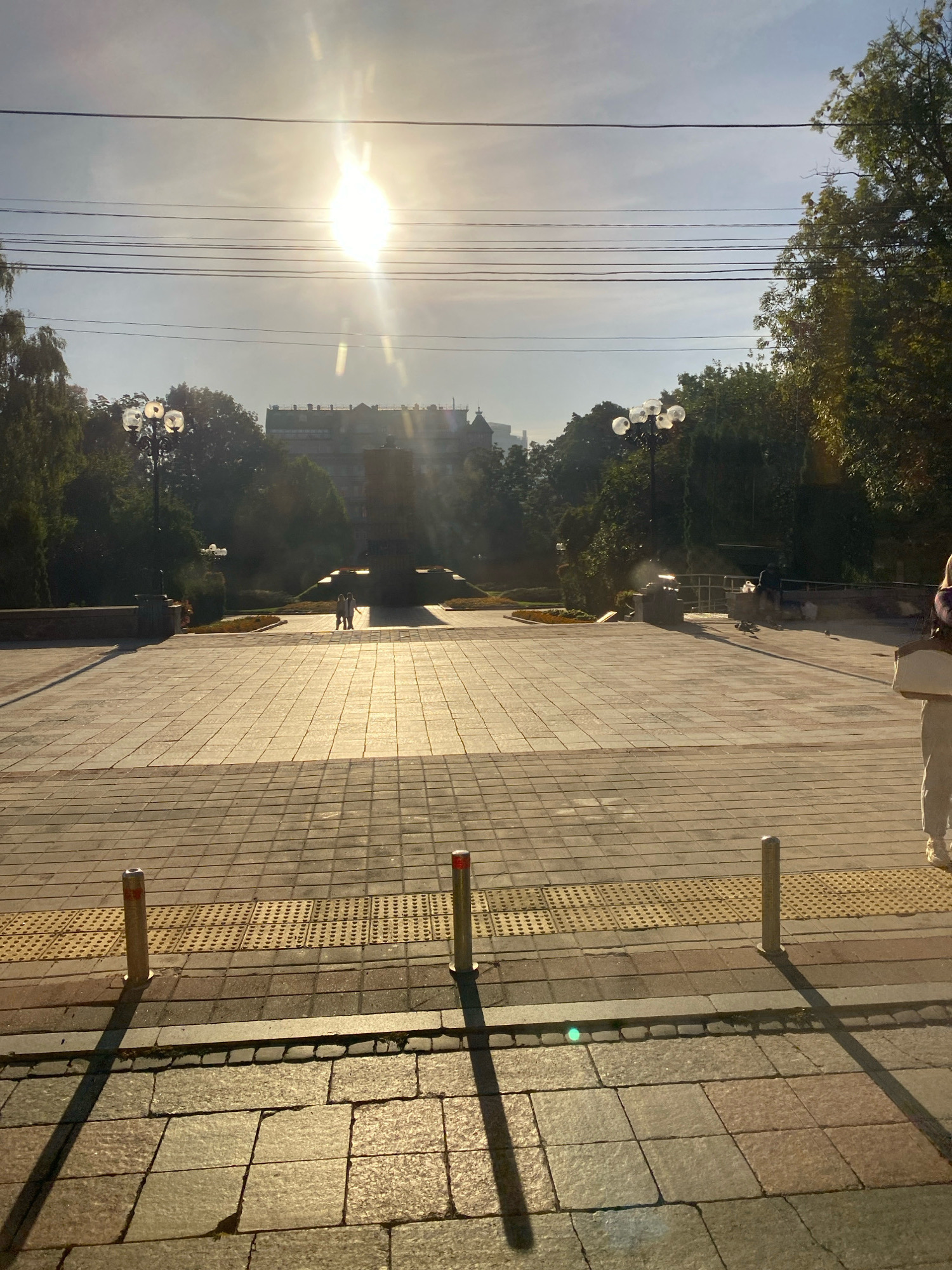 The image depicts a wide, sun-drenched pedestrian plaza paved with light-colored square tiles. The sun is high in the sky, casting long shadows and creating a bright glare. In the background, there is a large building with multiple stories and a row of lights along its roofline. Trees line the edges of the plaza, and several streetlights with multiple globes are visible. Two figures, seemingly a couple, are walking in the middle distance towards a monument or raised structure. Another person, dressed in light-colored pants and a white top, is standing on the right side of the frame, facing away from the camera and holding a flat, light-colored object. Several bollards are positioned along the edge of the plaza. The overall scene suggests a public outdoor space in a city, bathed in the warm light of the late afternoon or early morning sun.