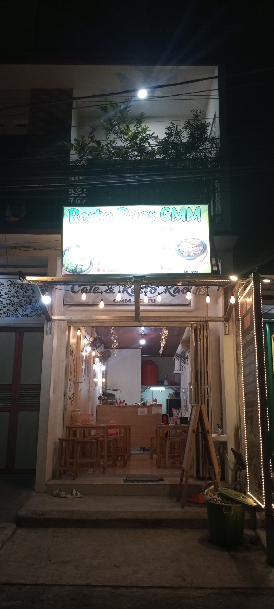 A well-lit restaurant sign, "Resto Paos GMM," is displayed above the entrance of a cafe and resto named "Cafe & Resto Kao" in the city of Setu, Indonesia. The sign features images of food and text indicating prices. The cafe entrance is open, revealing tables and chairs set up for dining. Inside, a counter is visible with appliances and what appears to be a water dispenser. Decorative lights are strung across the facade of the cafe, and a green trash can sits outside. The overall scene depicts a casual dining establishment at night.