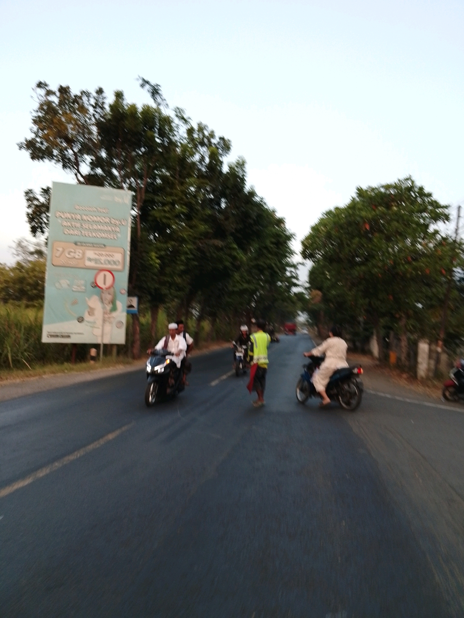 The image depicts a road in Pasuruan, Indonesia, with several motorcycles and pedestrians. On the left side of the road, a billboard advertises mobile phone data plans, featuring a price of Rp15,000 for 7 GB. A man wearing a white shirt and peci is riding a black motorcycle with another person behind him. Further down the road, a person in a bright yellow vest and red sash is standing near a motorcycle. On the right side, a person in a light-colored outfit is riding a motorcycle. The scene appears to be during dusk, with trees lining the road. The overall impression is one of everyday street life in Indonesia, with traffic and commercial advertising integrated into the environment.