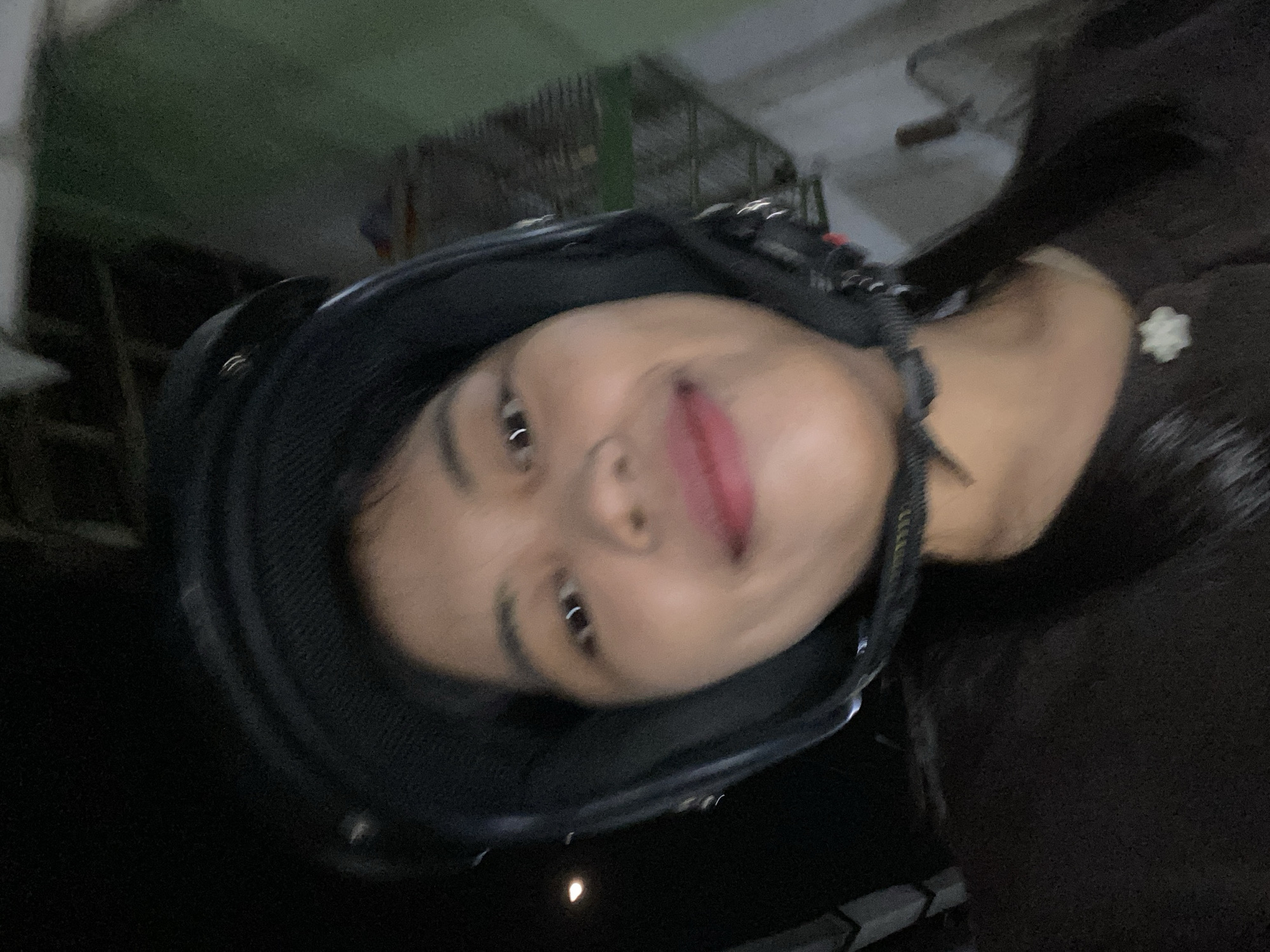 The image shows a close-up, Dutch angle selfie of a person wearing a black helmet. The person has dark hair and is looking towards the camera, with their tongue slightly sticking out. Their lips are painted with a vibrant pink lipstick. The background is dark and out of focus, with some faint hints of what appear to be structures or vehicles. The lighting is dim, suggesting it might be nighttime or indoors. There is no visible text in the image. The overall impression is casual and playful. The context of Madiun, Indonesia, does not provide specific visual cues within the image itself.